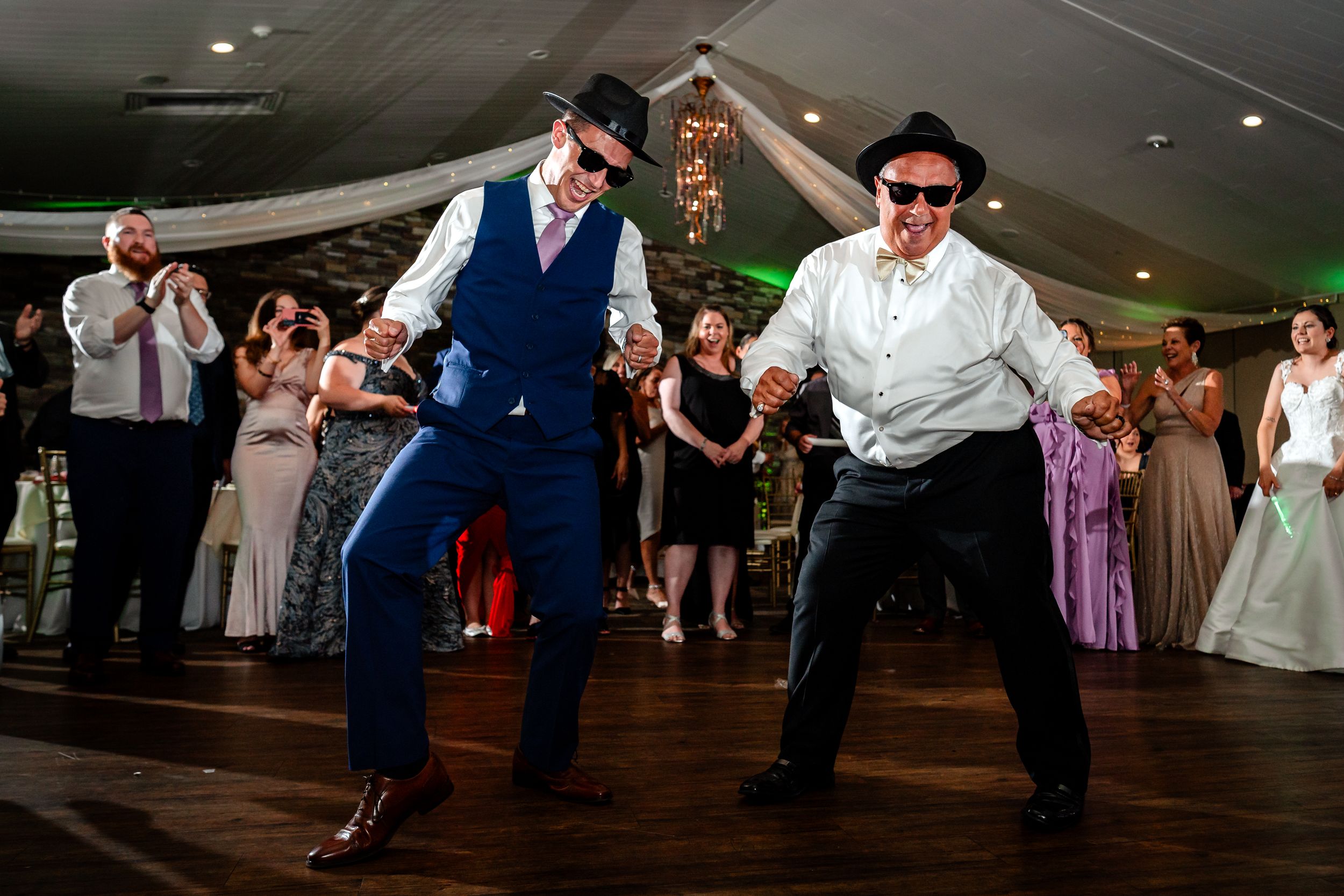 Wedding guests dancing and jumping energetically on a dark dance floor with ambient lighting.