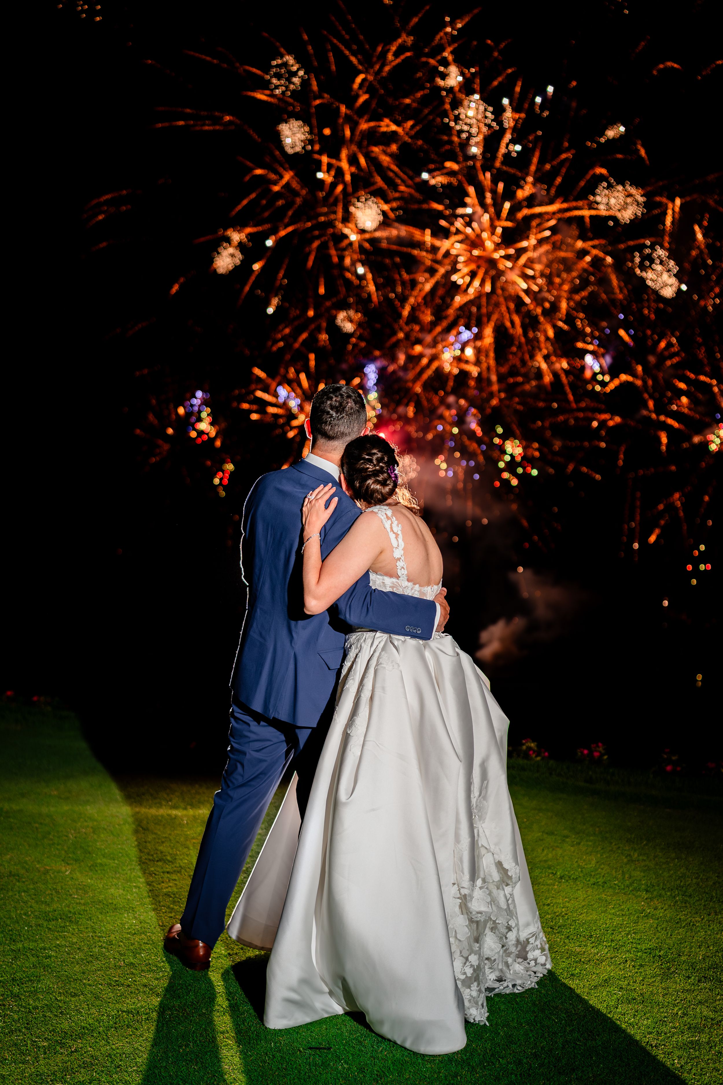 Couple watching fireworks display during their wedding celebration on a dark grassy lawn.