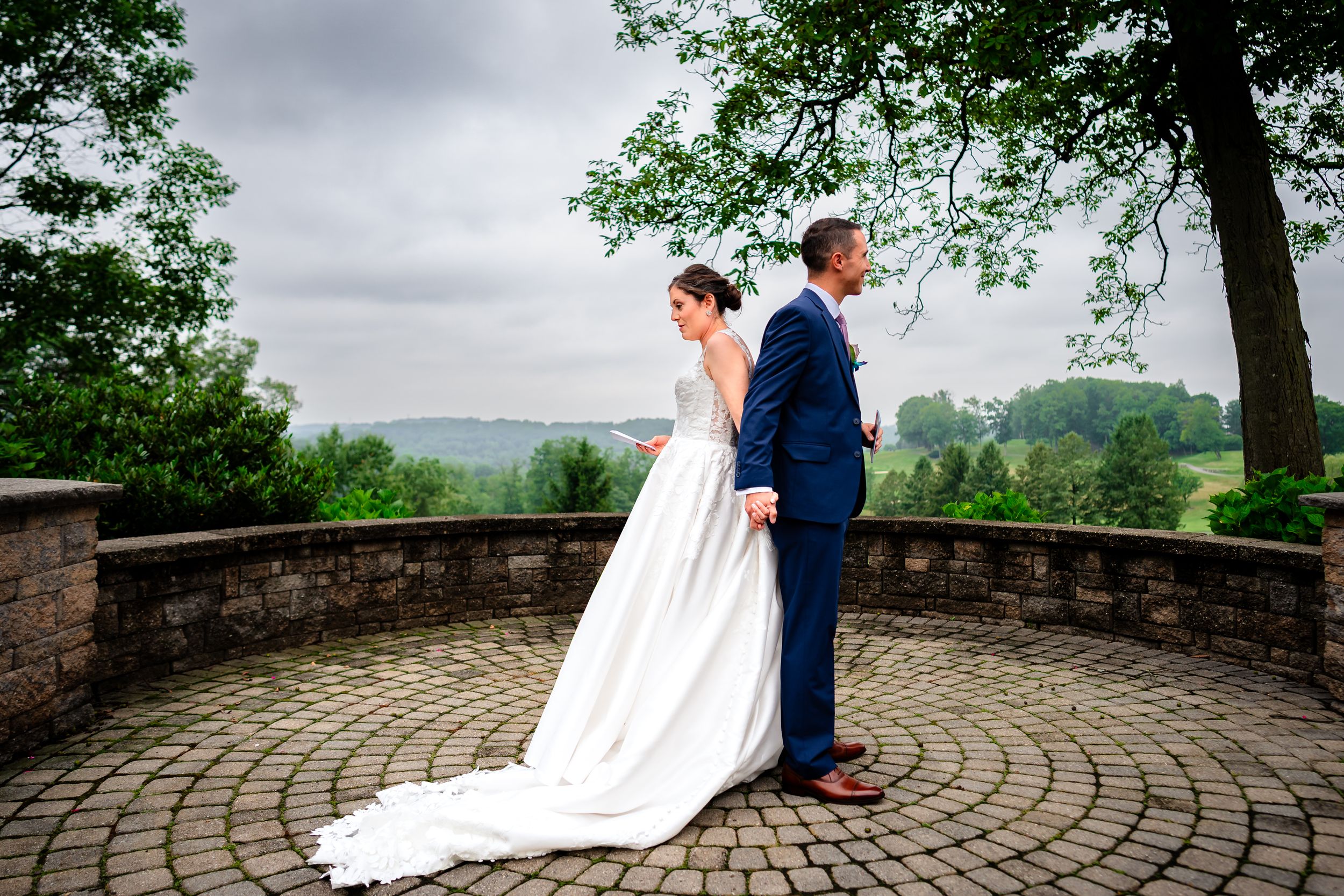 A couple in wedding attire embraces on a circular stone patio overlooking a scenic mountain landscape.