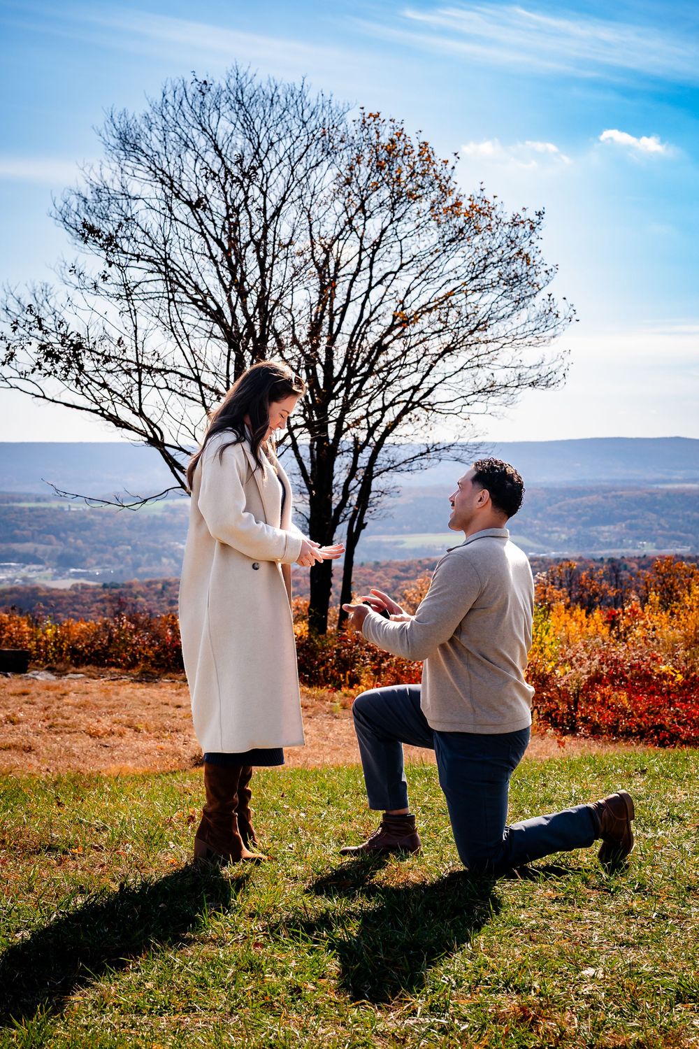 A couple shares a romantic proposal moment on a hilltop during autumn with a bare tree and colorful fall foliage in background.