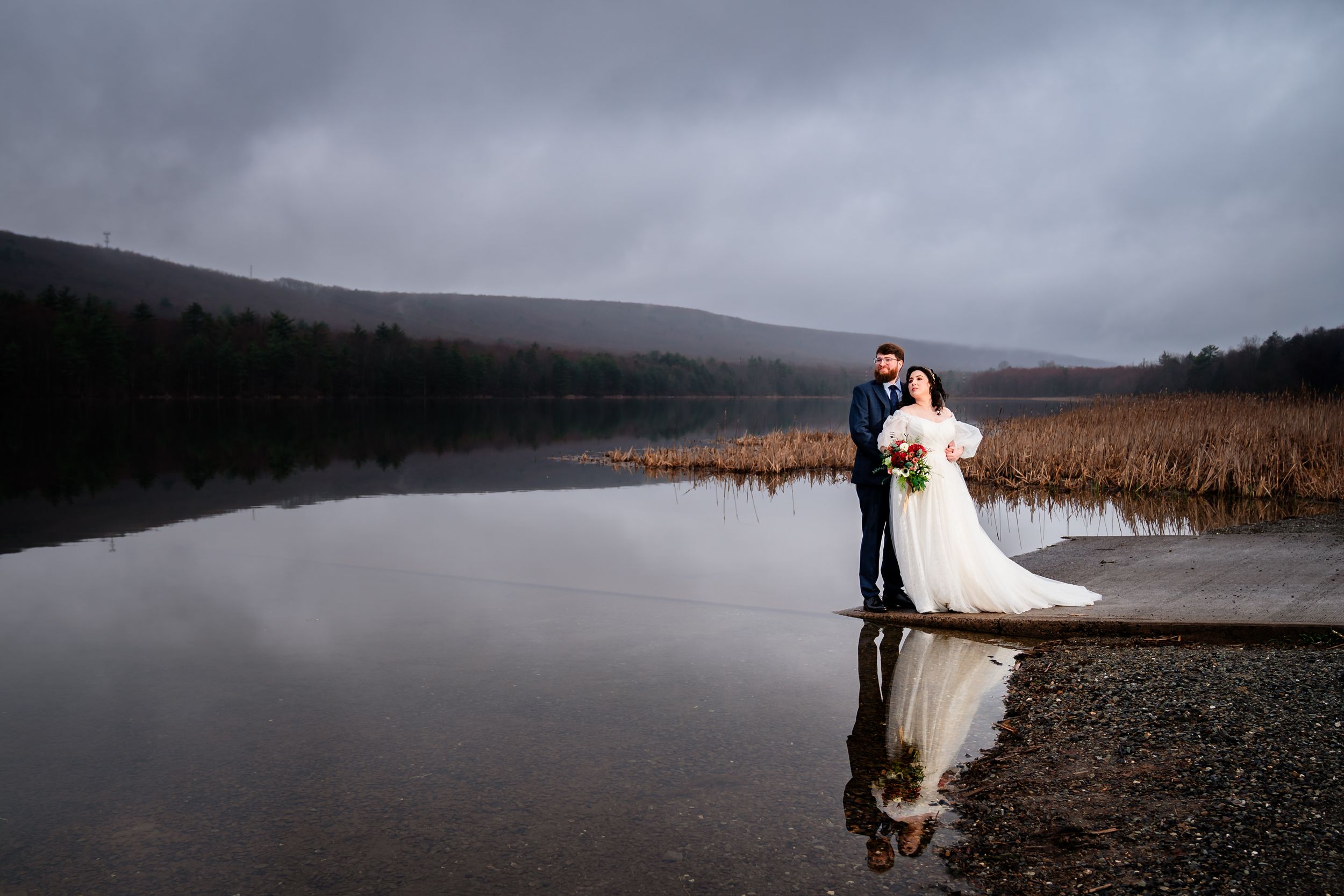A romantic wedding photo session on a misty lake shore with dramatic cloudy skies and tall reeds in the background.