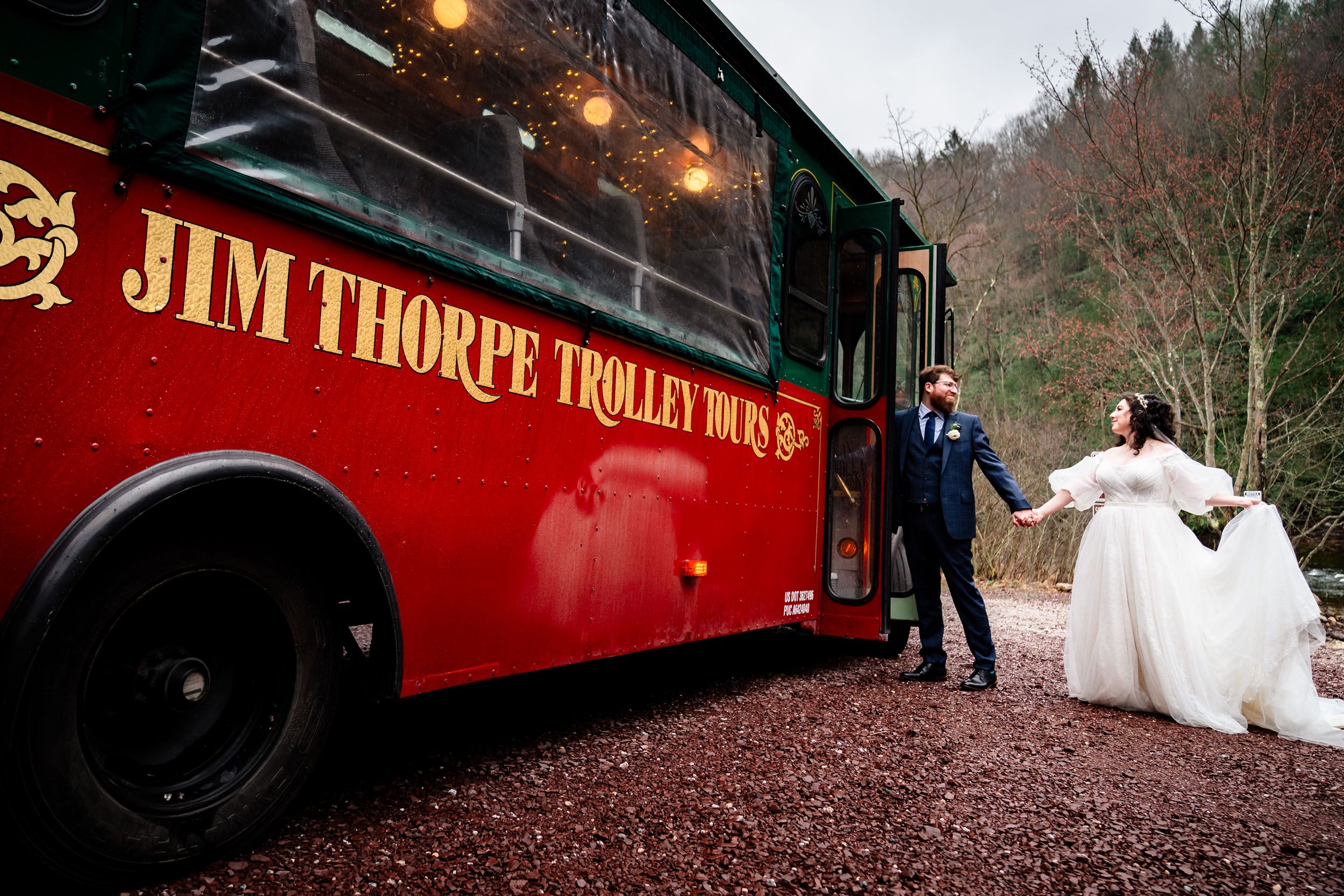 A red vintage train car decorated for a wedding celebration with guests and string lights in a wooded setting.