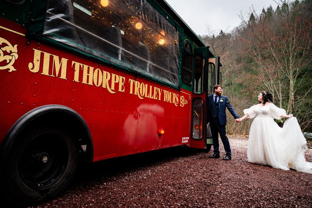 A red vintage train car decorated for a wedding celebration with guests and string lights in a wooded setting.