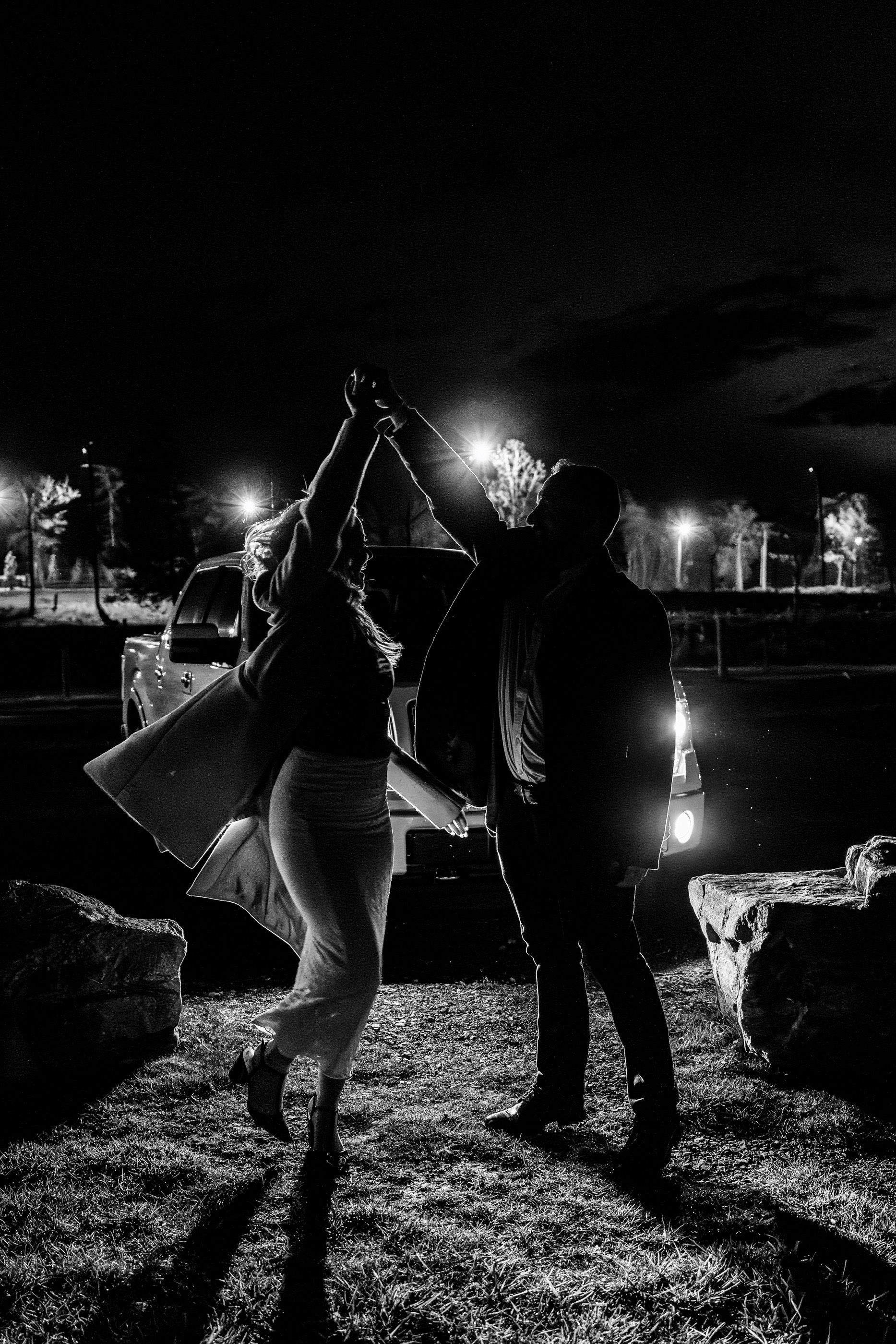 A black and white nighttime photo of two people dancing together under street lights in a park.