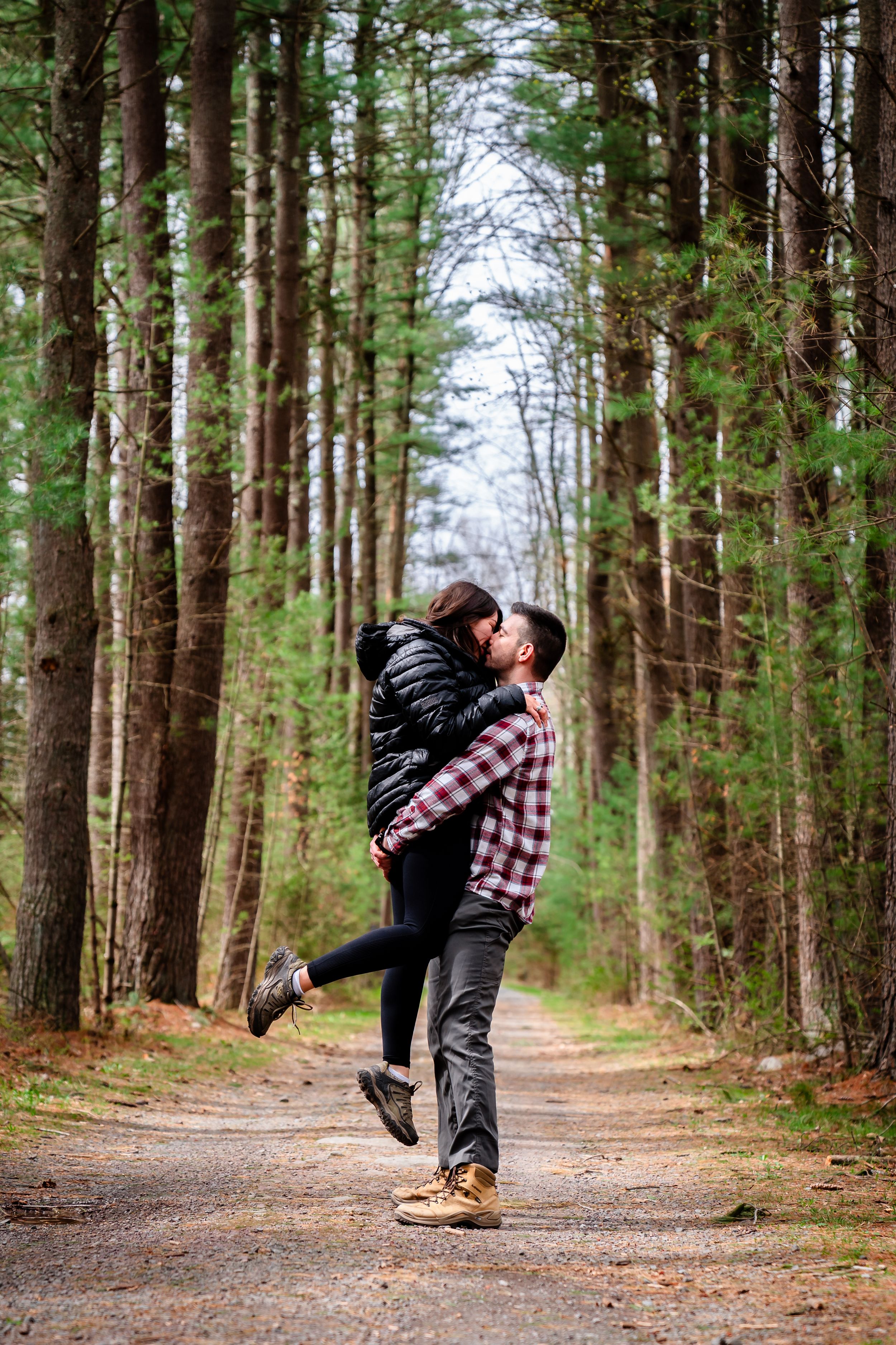 A couple shares a romantic kiss while hiking on a tree-lined forest trail during autumn.