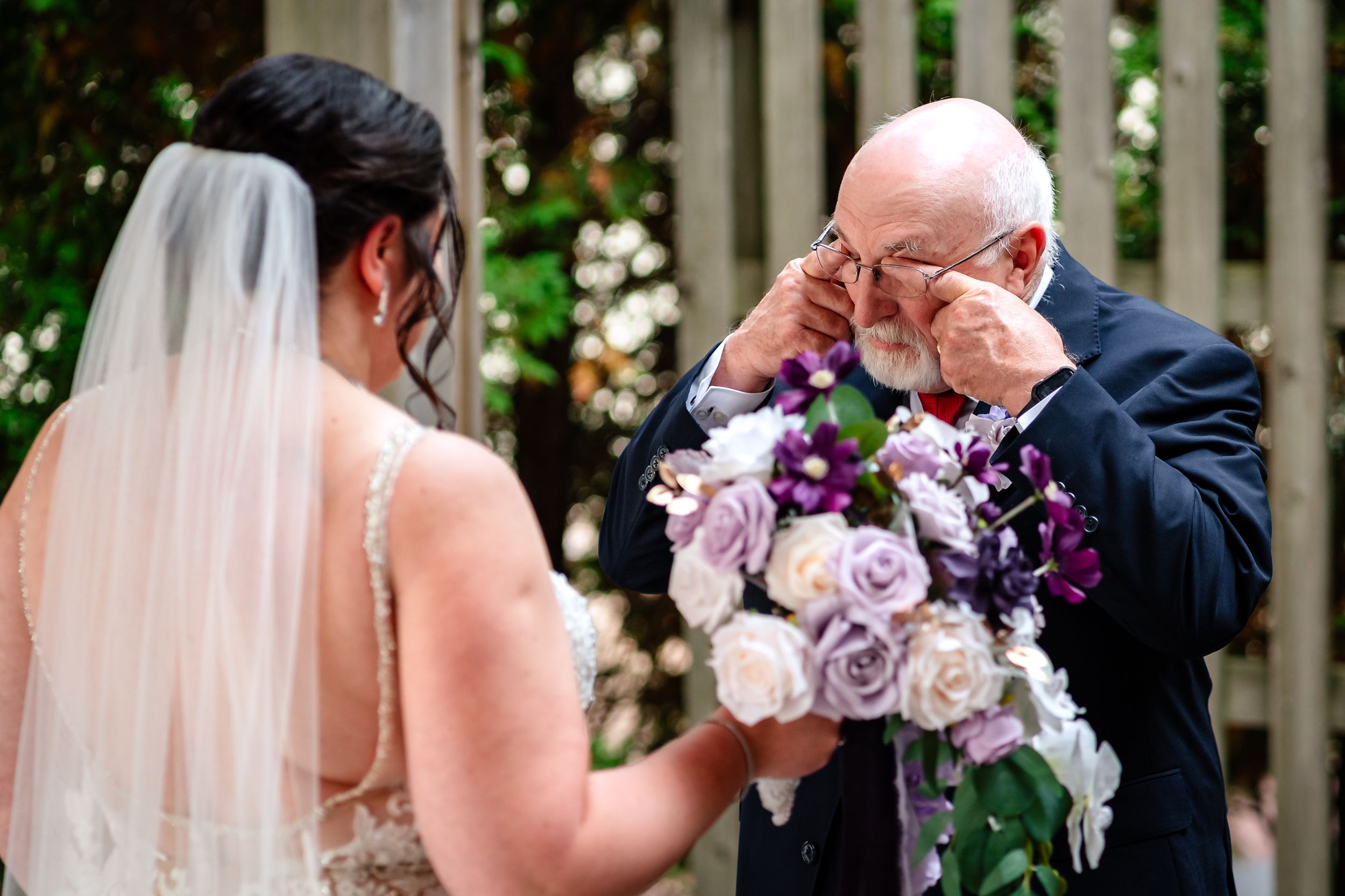 A wedding couple holds purple and pink bouquets while standing in front of a white picket fence with greenery.