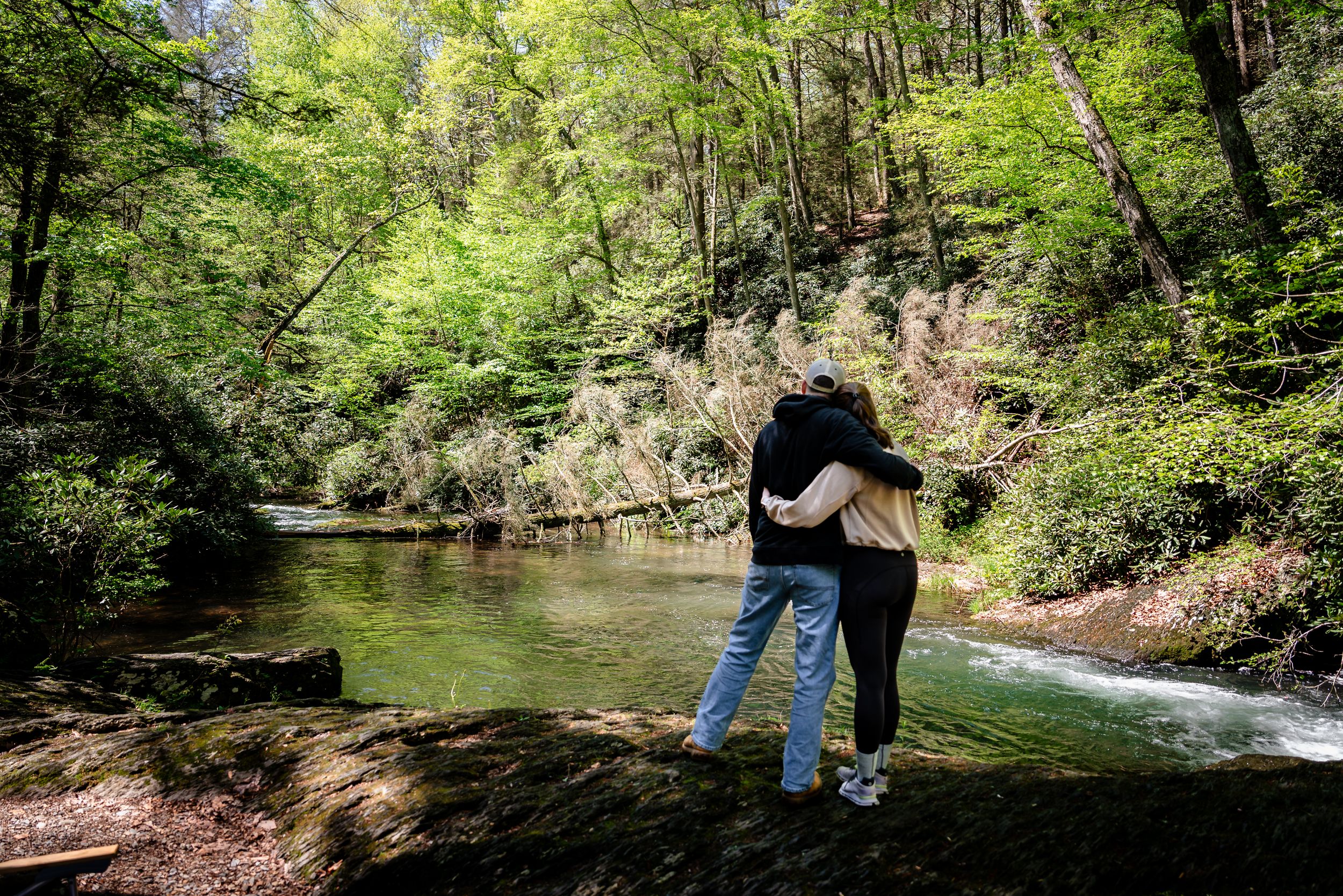 Two people walk along a scenic wooded trail beside a flowing stream on a sunny day.