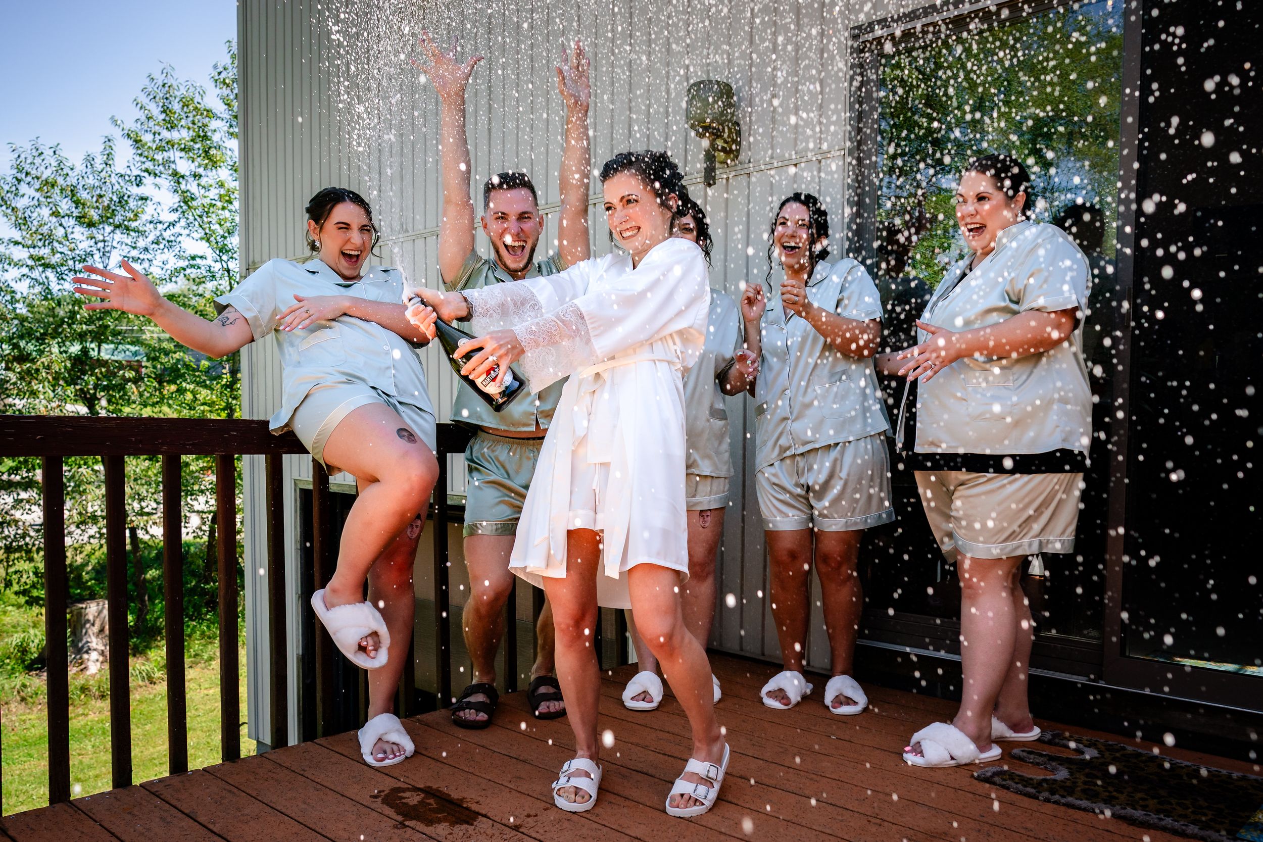 Groomsmen in white shirts and khaki shorts sit casually on a wooden porch railing outdoors.