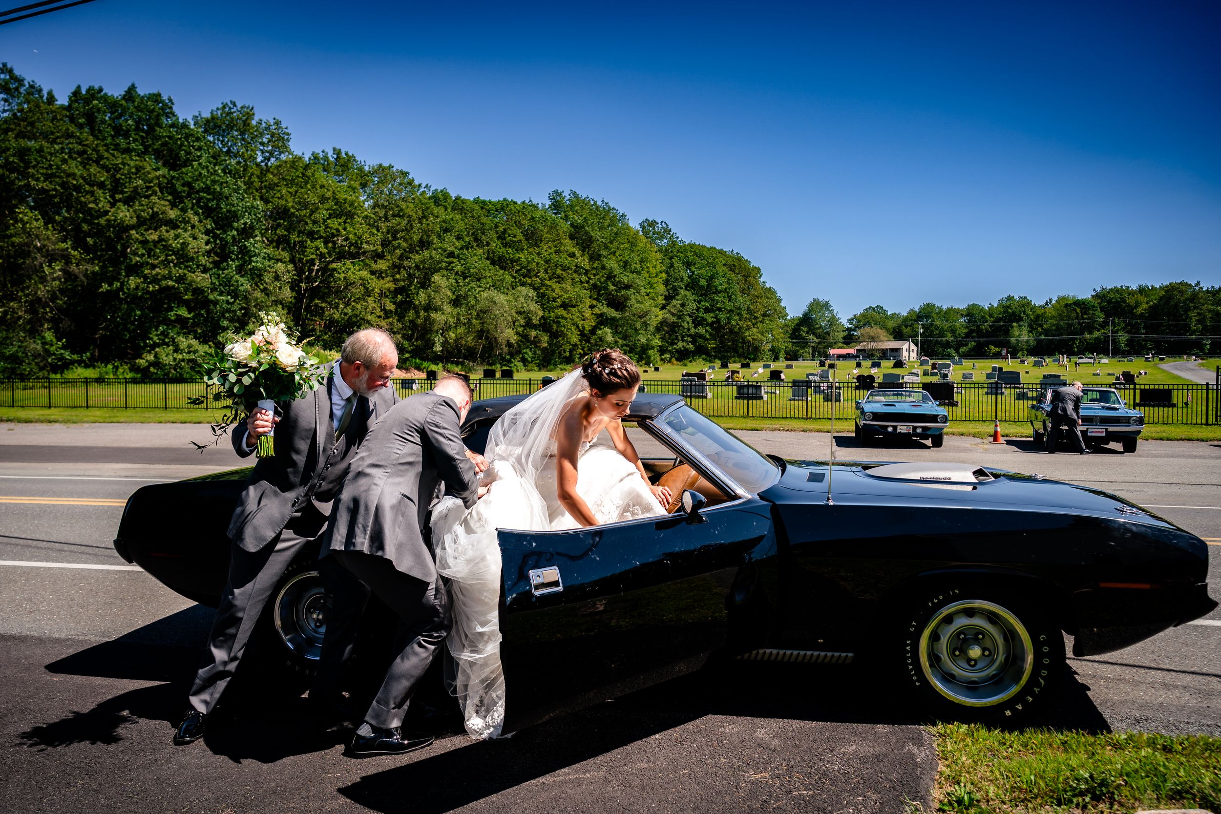 A classic black sports car is pushed by several people across a sunny field on a beautiful summer day.