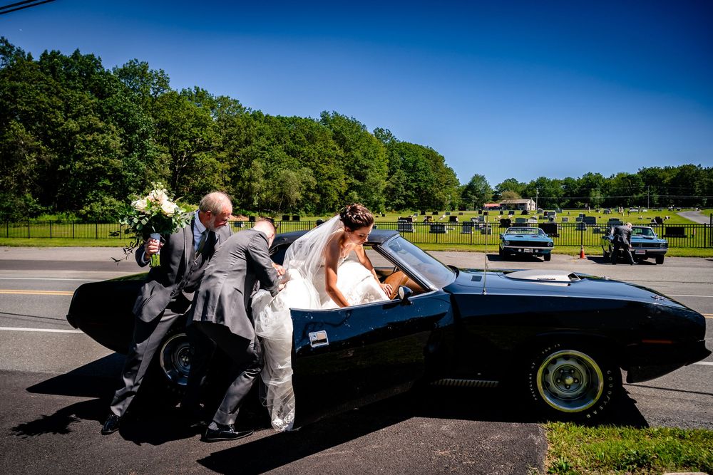 A classic black sports car is pushed by several people across a sunny field on a beautiful summer day.