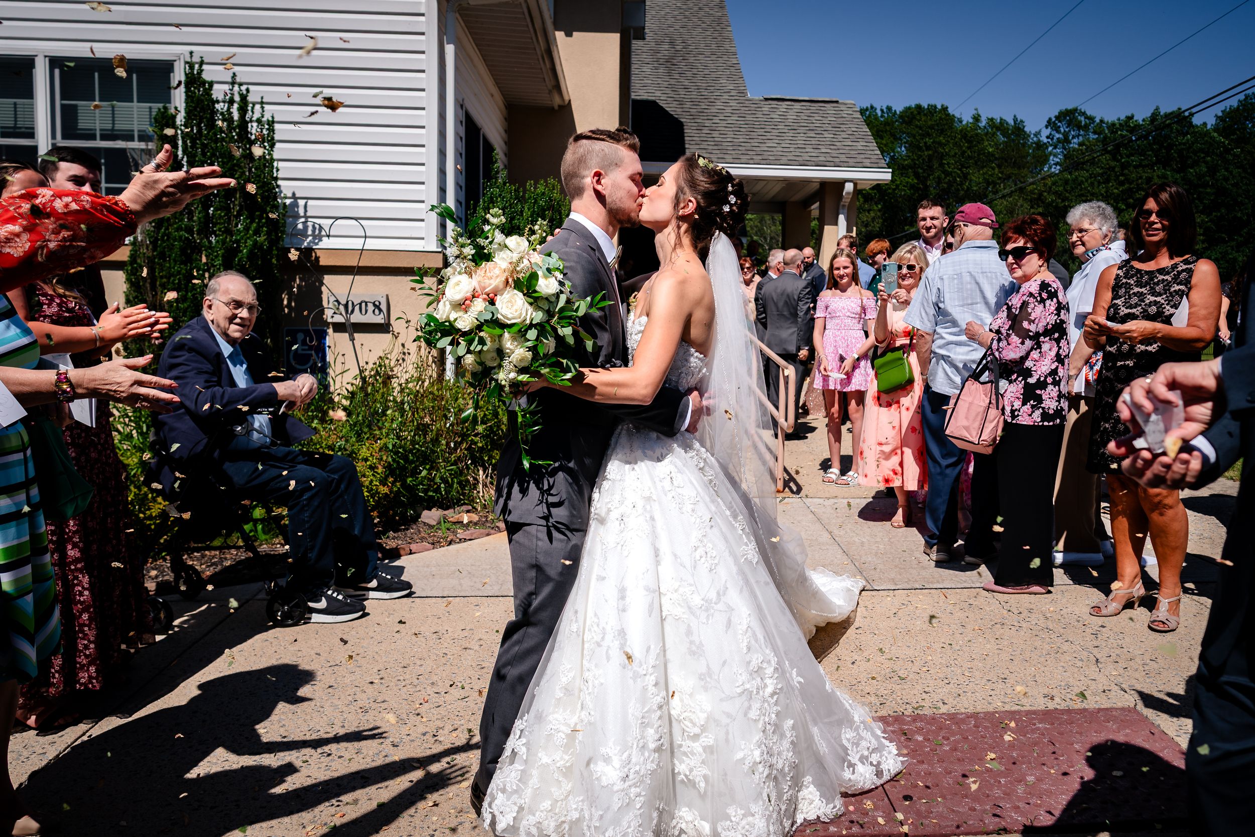Guests shower rose petals on a couple during an outdoor wedding celebration on a sunny day.