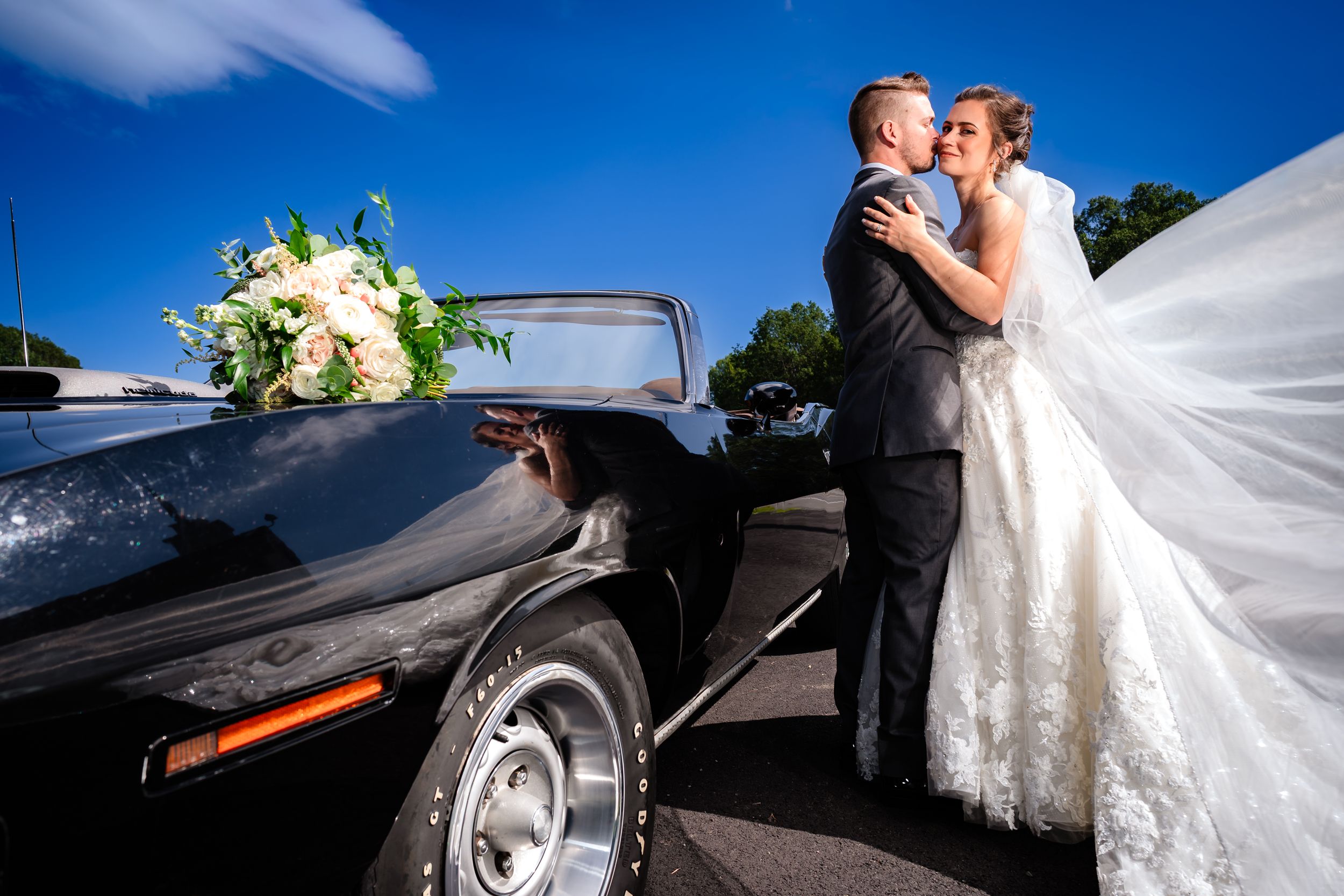 A white wedding dress flows elegantly next to a classic black car during an outdoor celebration.