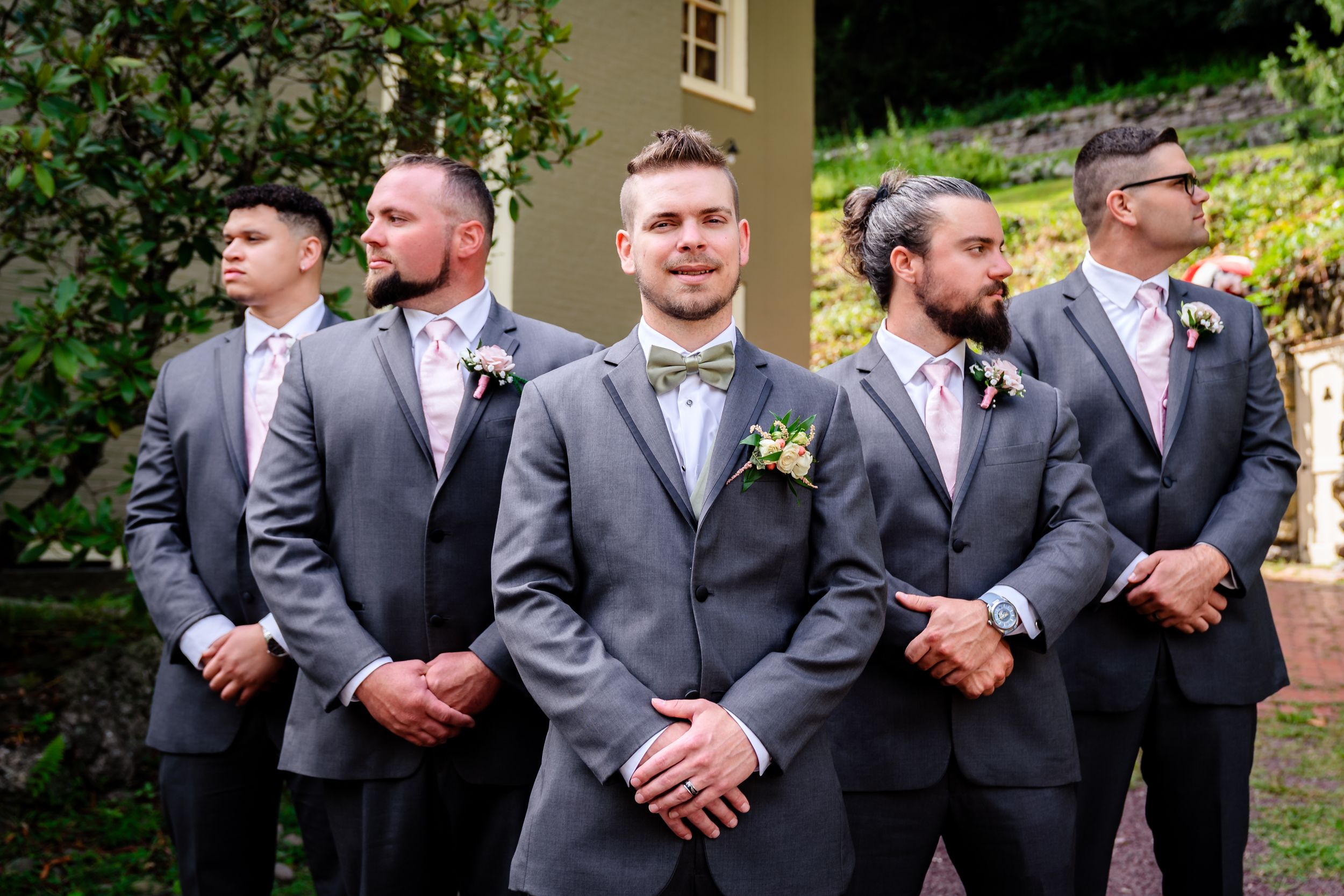 A row of groomsmen in matching gray suits stand together at an outdoor wedding venue.