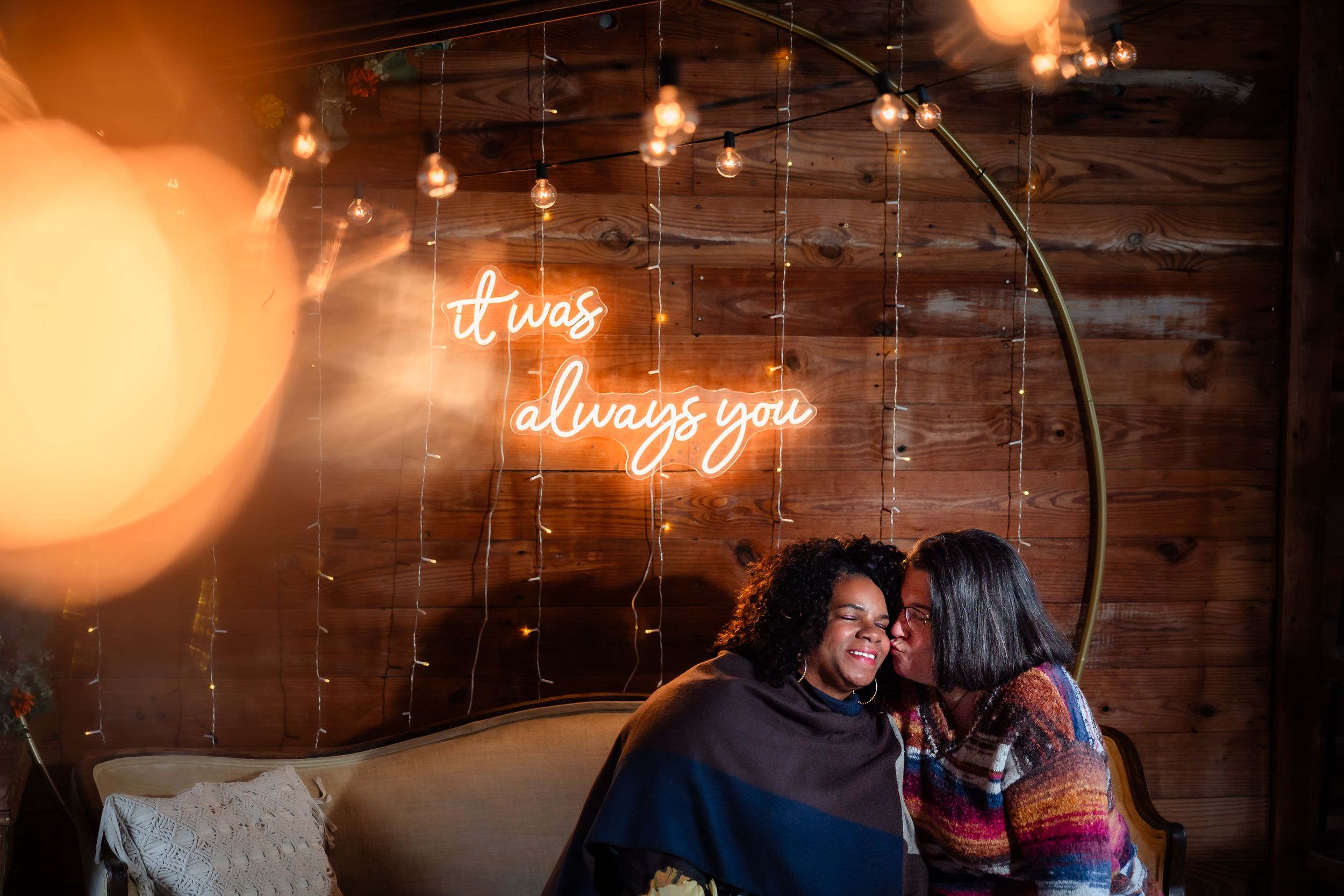 Romantic couple embraces under warm neon lighting and string lights against rustic wood wall.