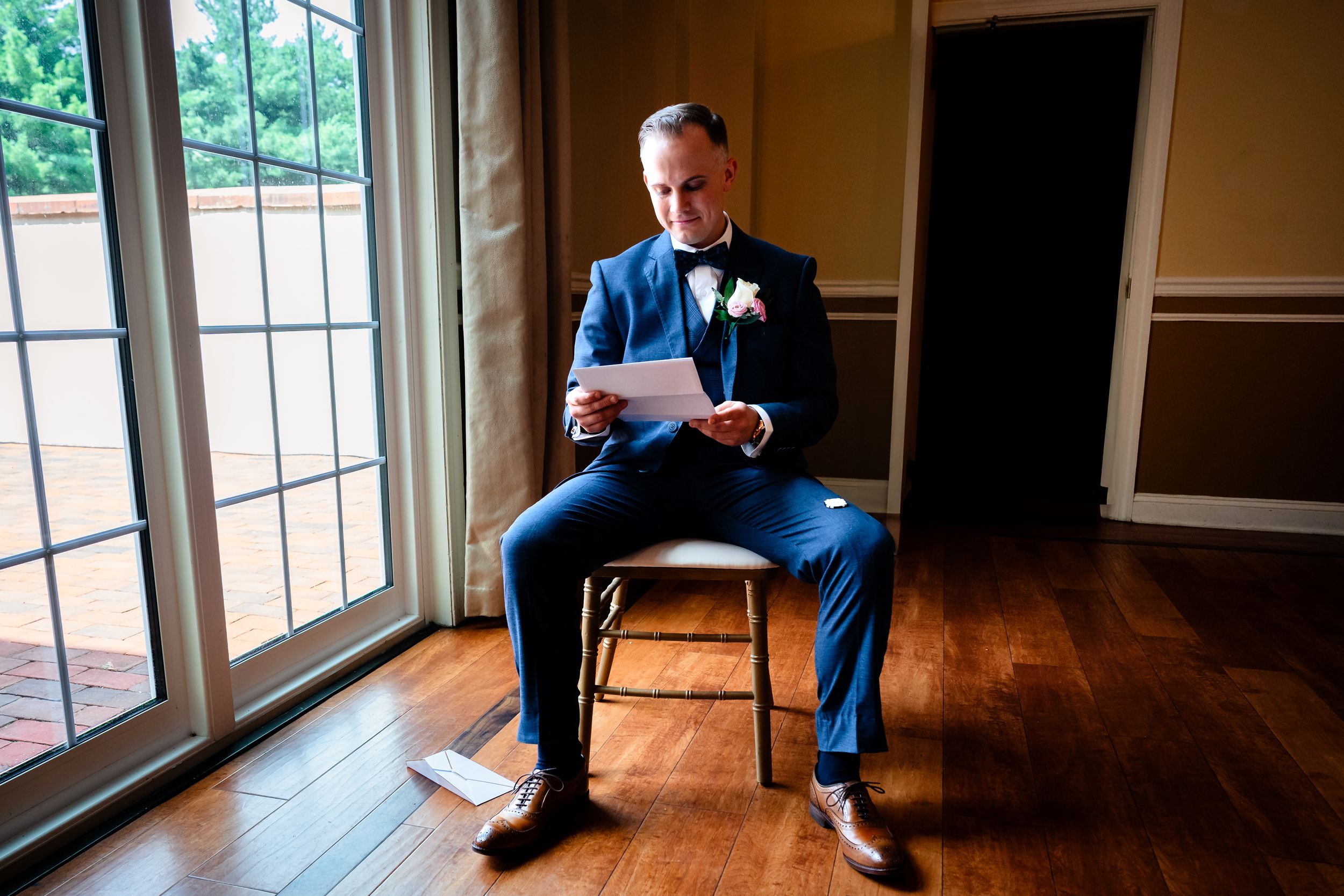 Person in a blue suit sits by window in an elegant room with hardwood floors and natural lighting.