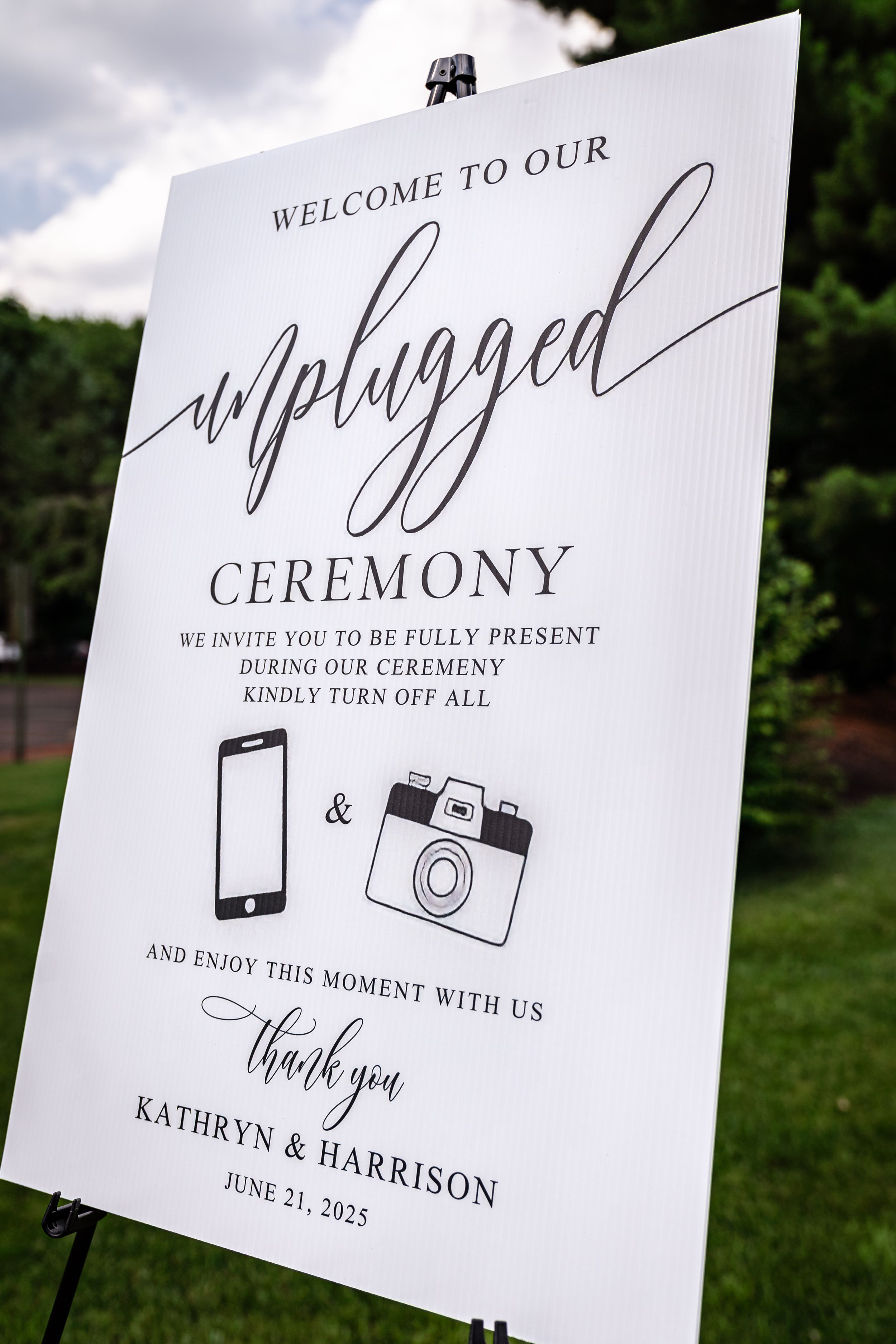 Wedding ceremony signage with elegant calligraphy and camera icons displayed outdoors against a natural background.