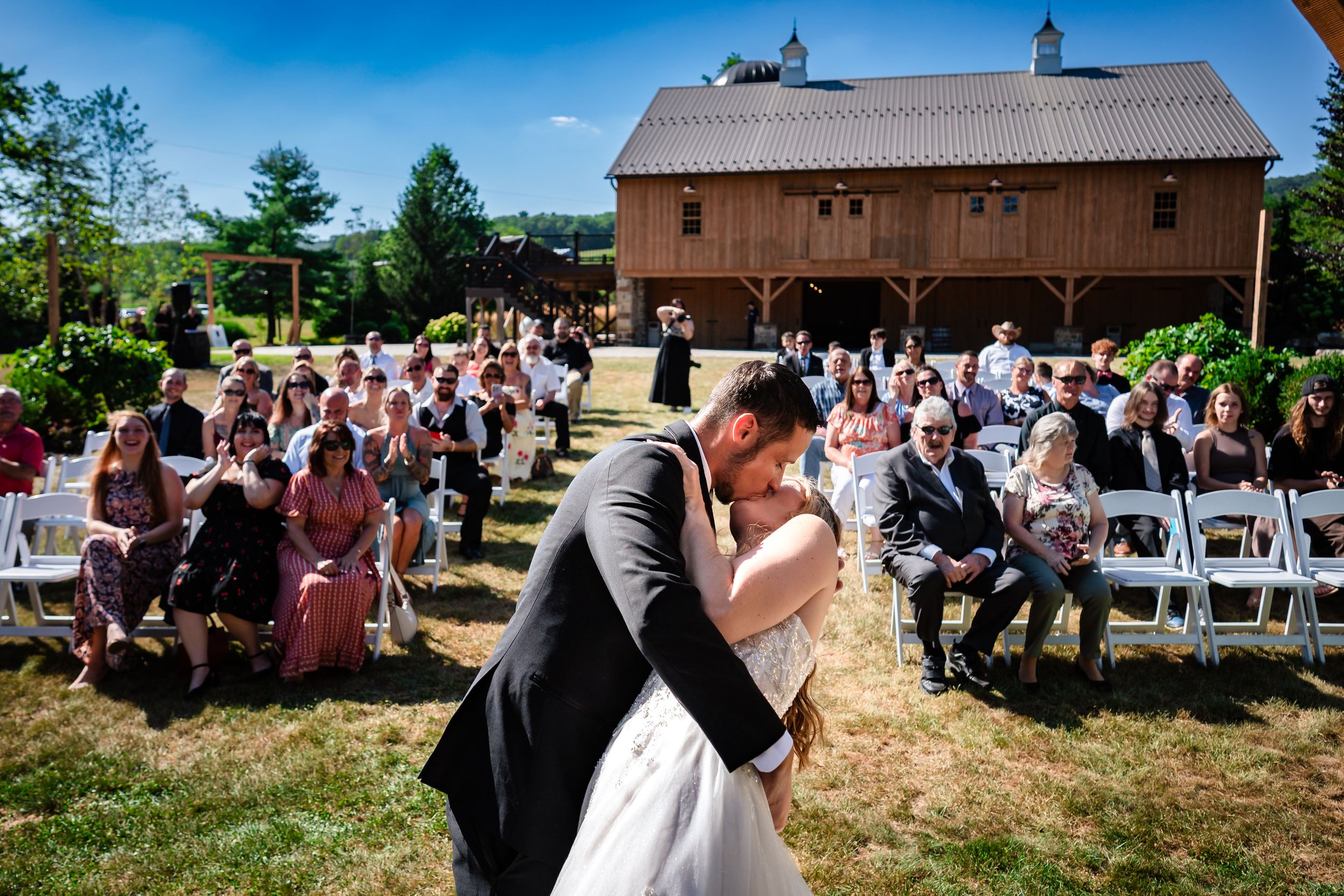 A romantic wedding ceremony takes place outside a rustic brown barn with guests seated on white chairs in a grassy field.