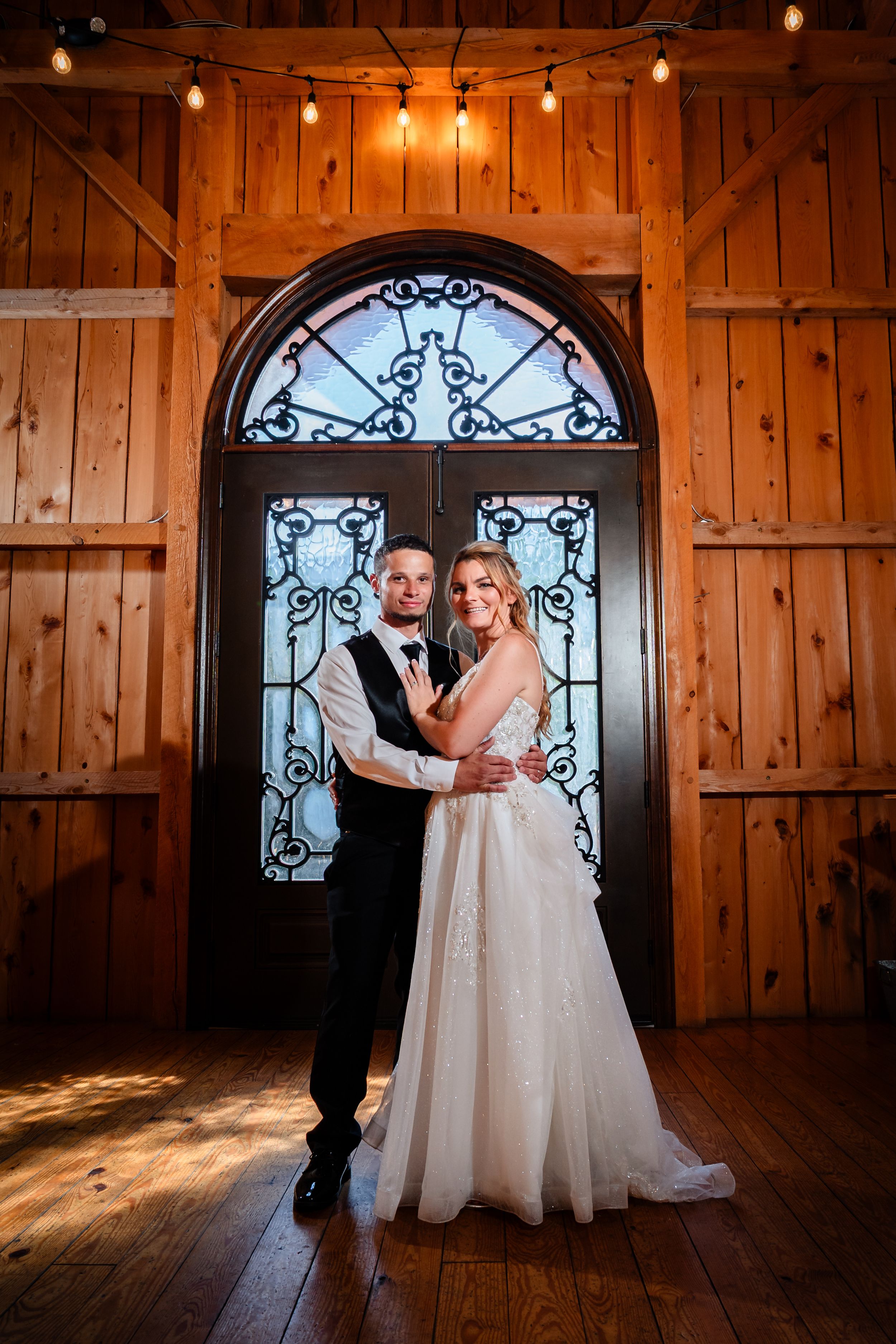 A romantic wedding couple dances together in a rustic wooden barn venue with decorative iron windows and warm lighting.