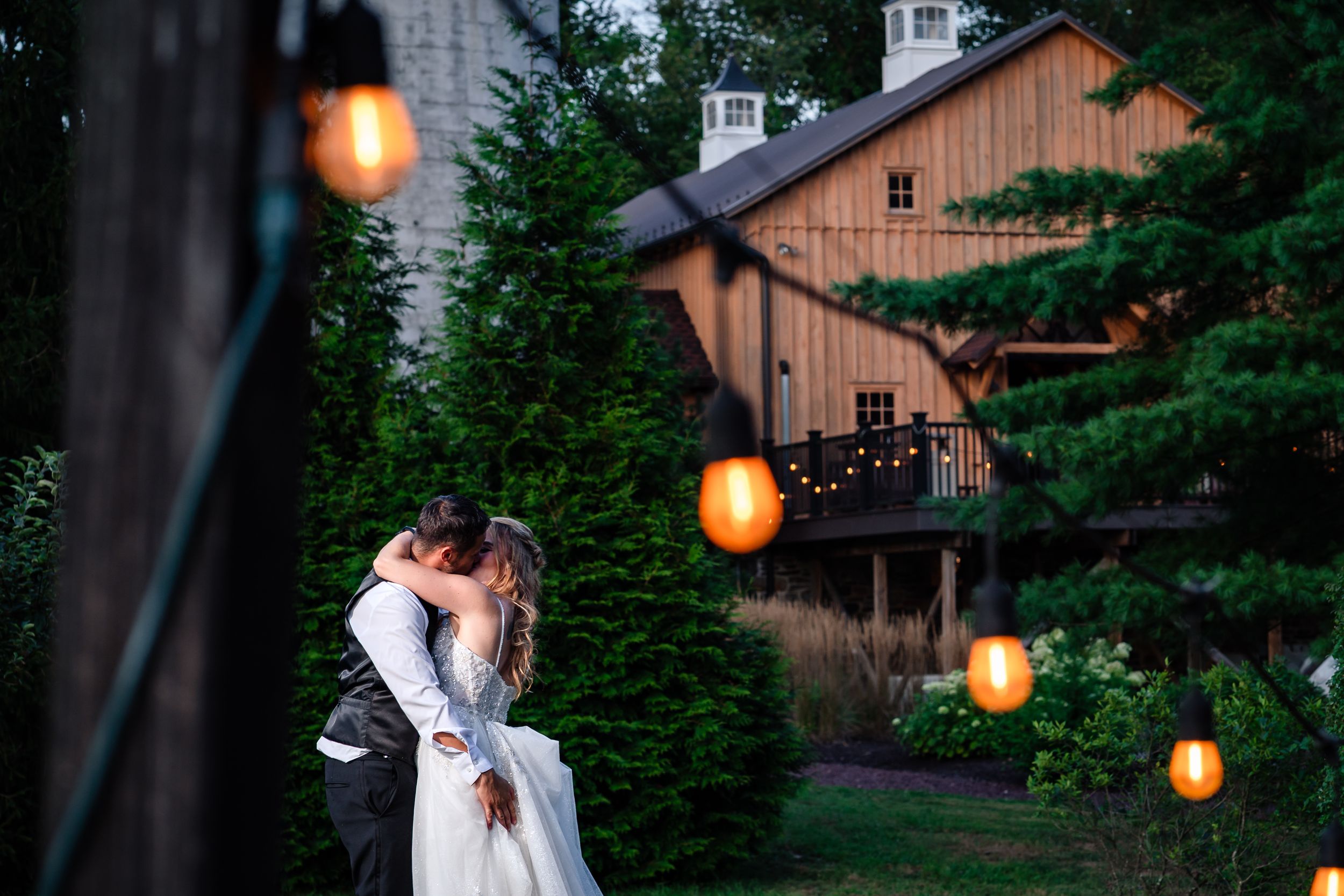 String lights illuminate a rustic outdoor wedding venue with wooden buildings and lush greenery at dusk.