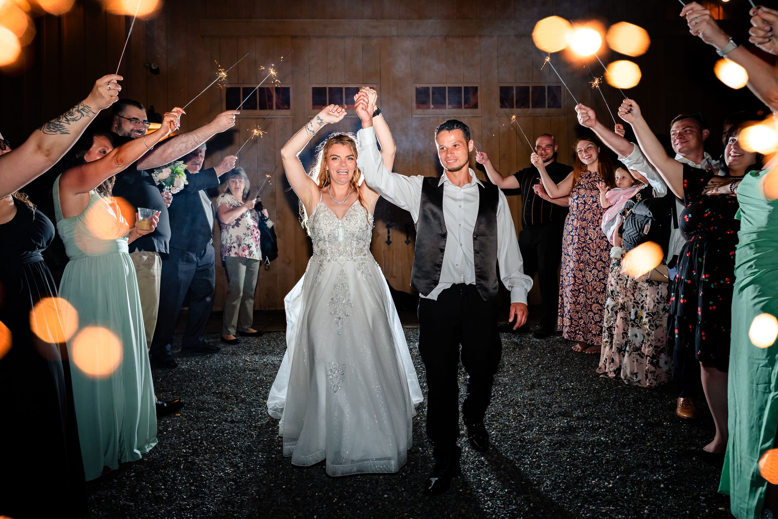 Newlyweds celebrate with guests during nighttime reception as sparklers illuminate their joyful exit along pathway.