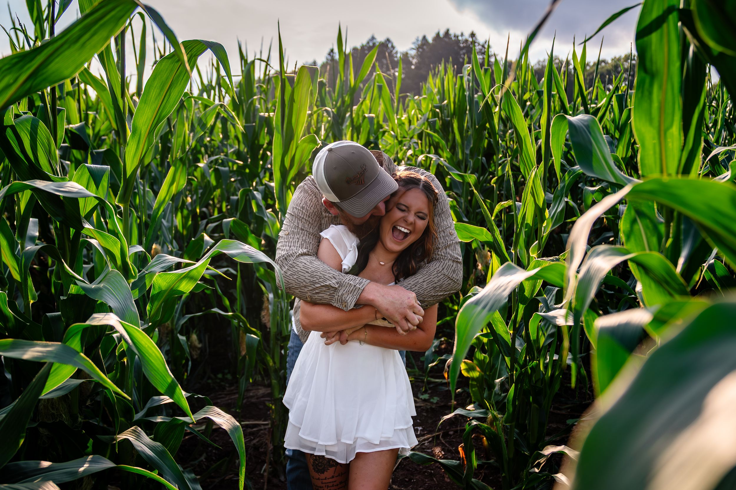 A couple embraces lovingly among tall green corn stalks in a summer field during a romantic photoshoot.
