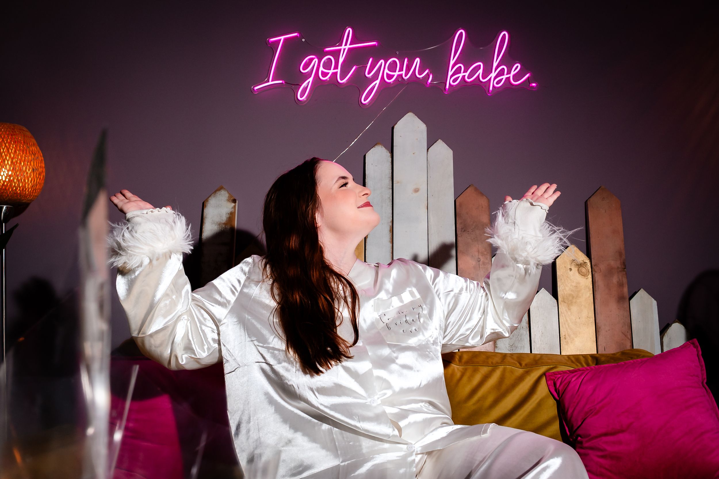 A person in a white satin robe lounges on pink furniture in a dimly lit room.