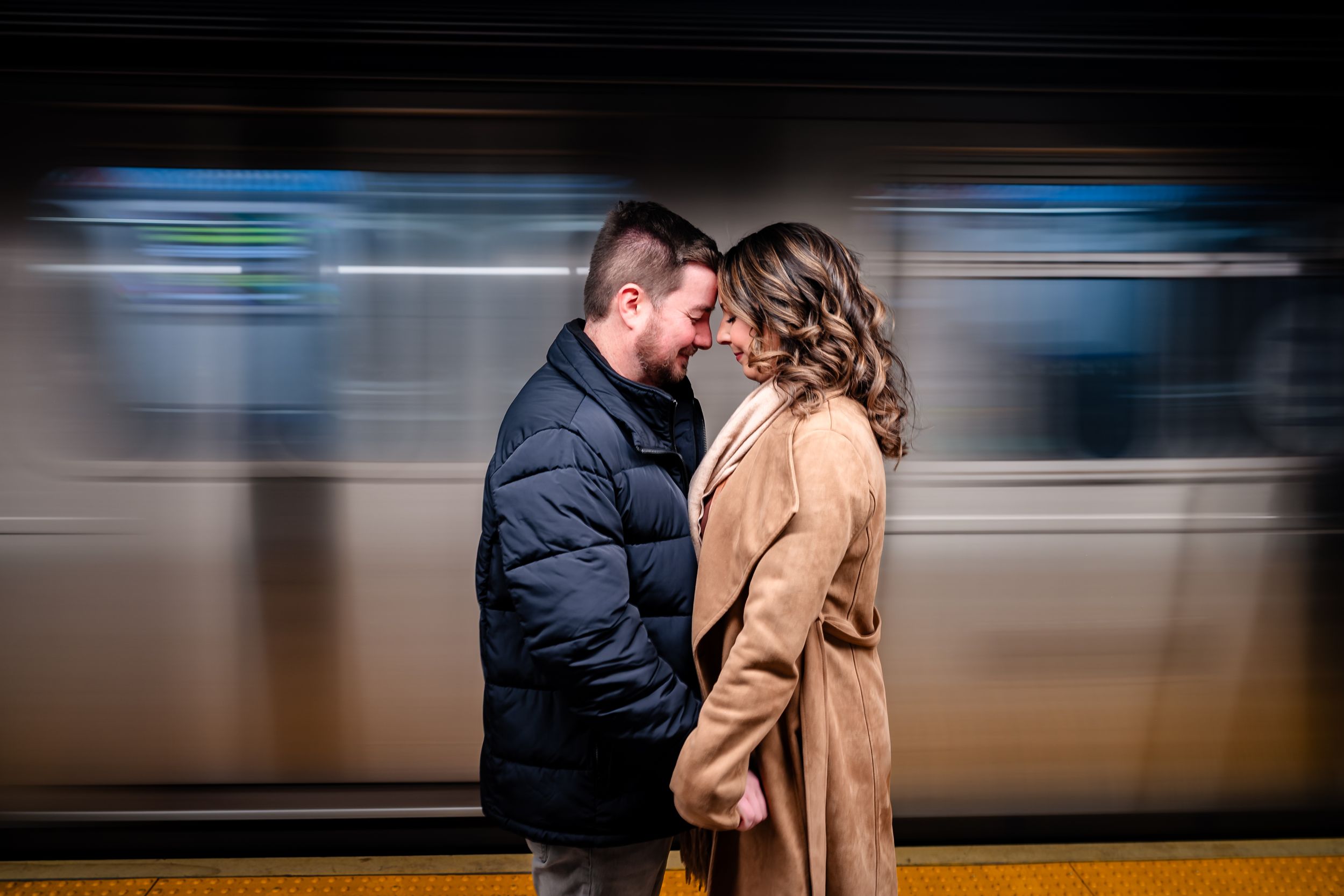 Two people embrace on a subway platform as trains pass by creating motion blur in the background.