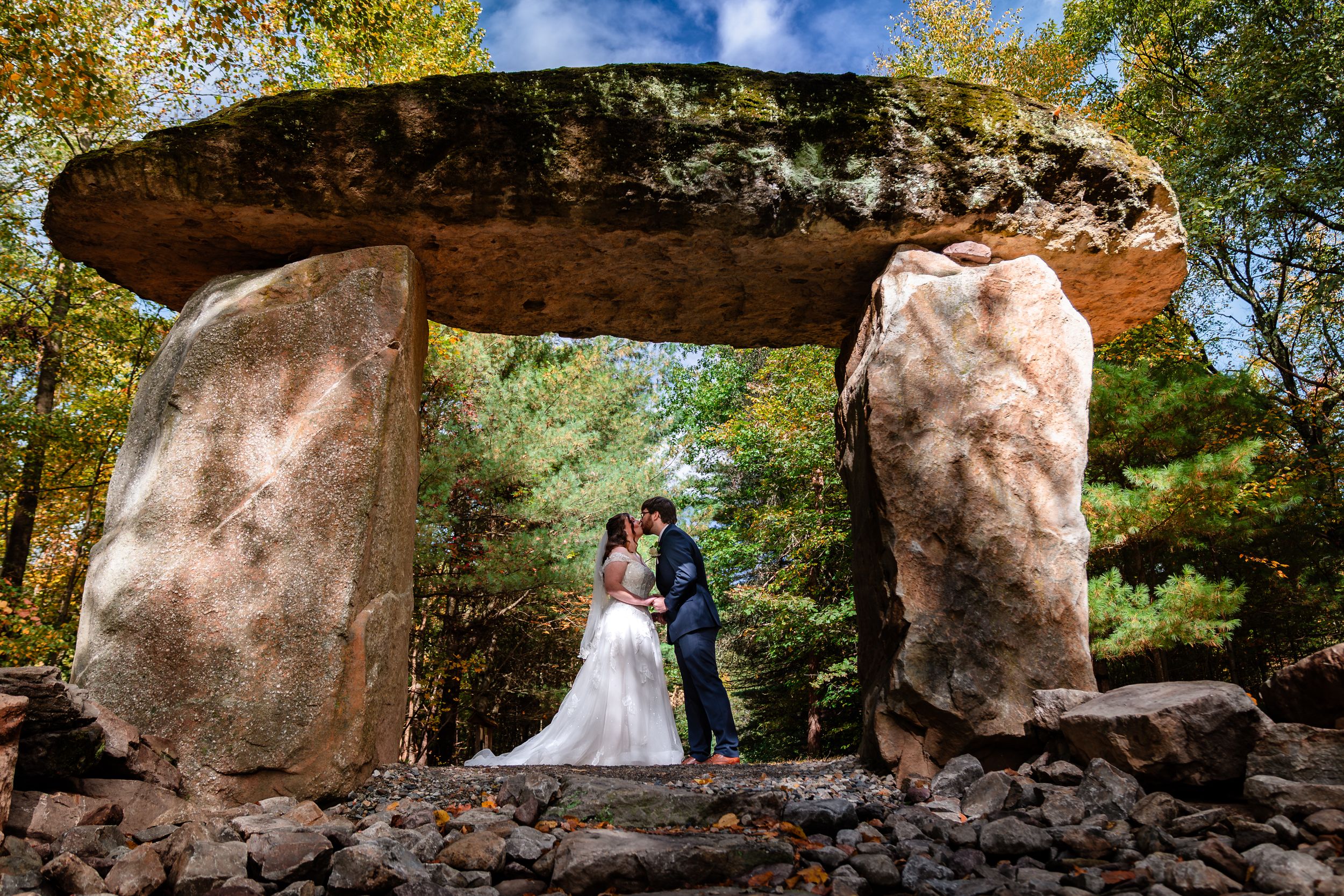 A romantic wedding photo session captured under natural rock formations in a scenic outdoor setting with autumn foliage.