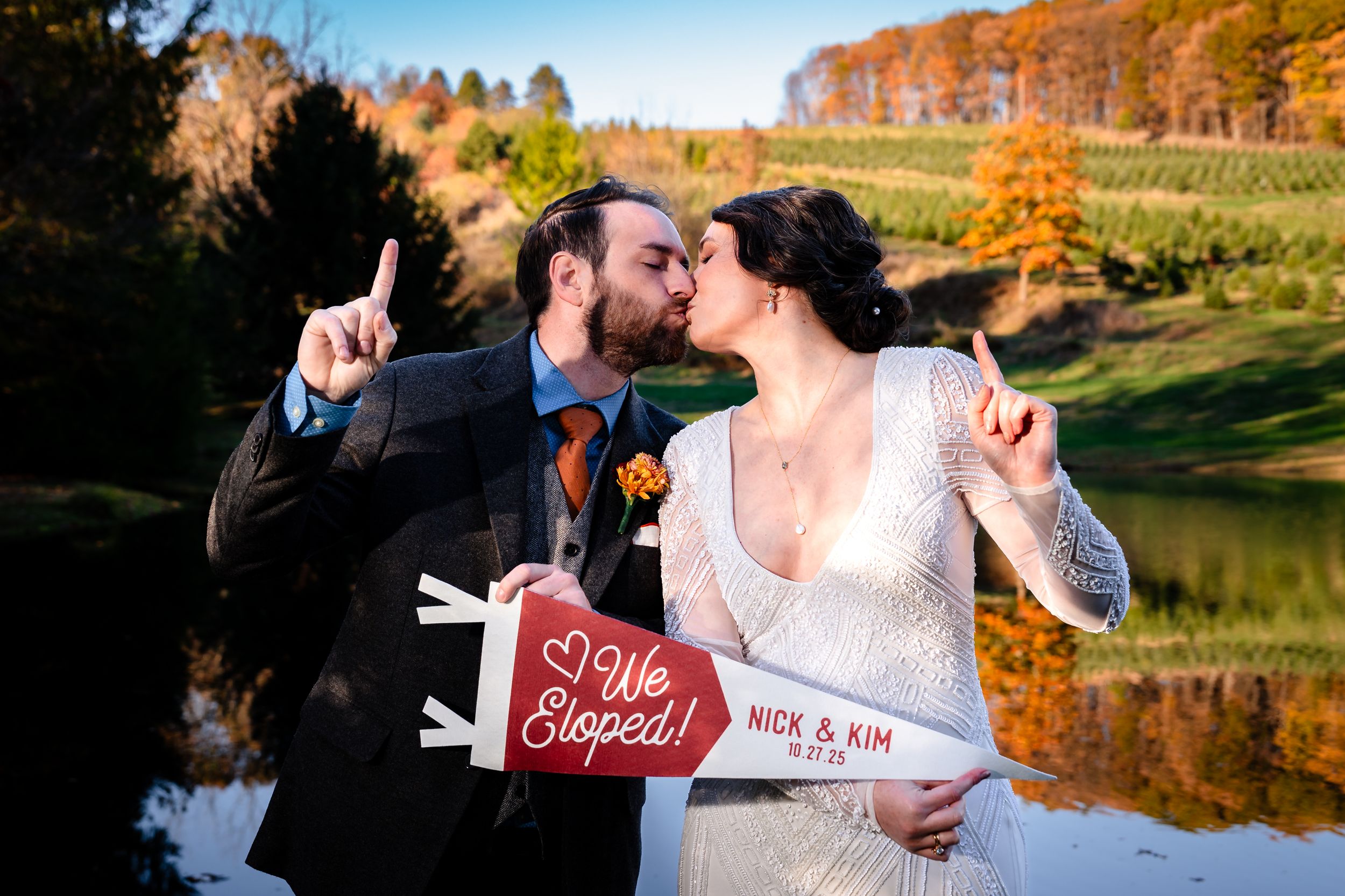 Romantic fall wedding photos captured by a lake with vibrant autumn foliage in the background.