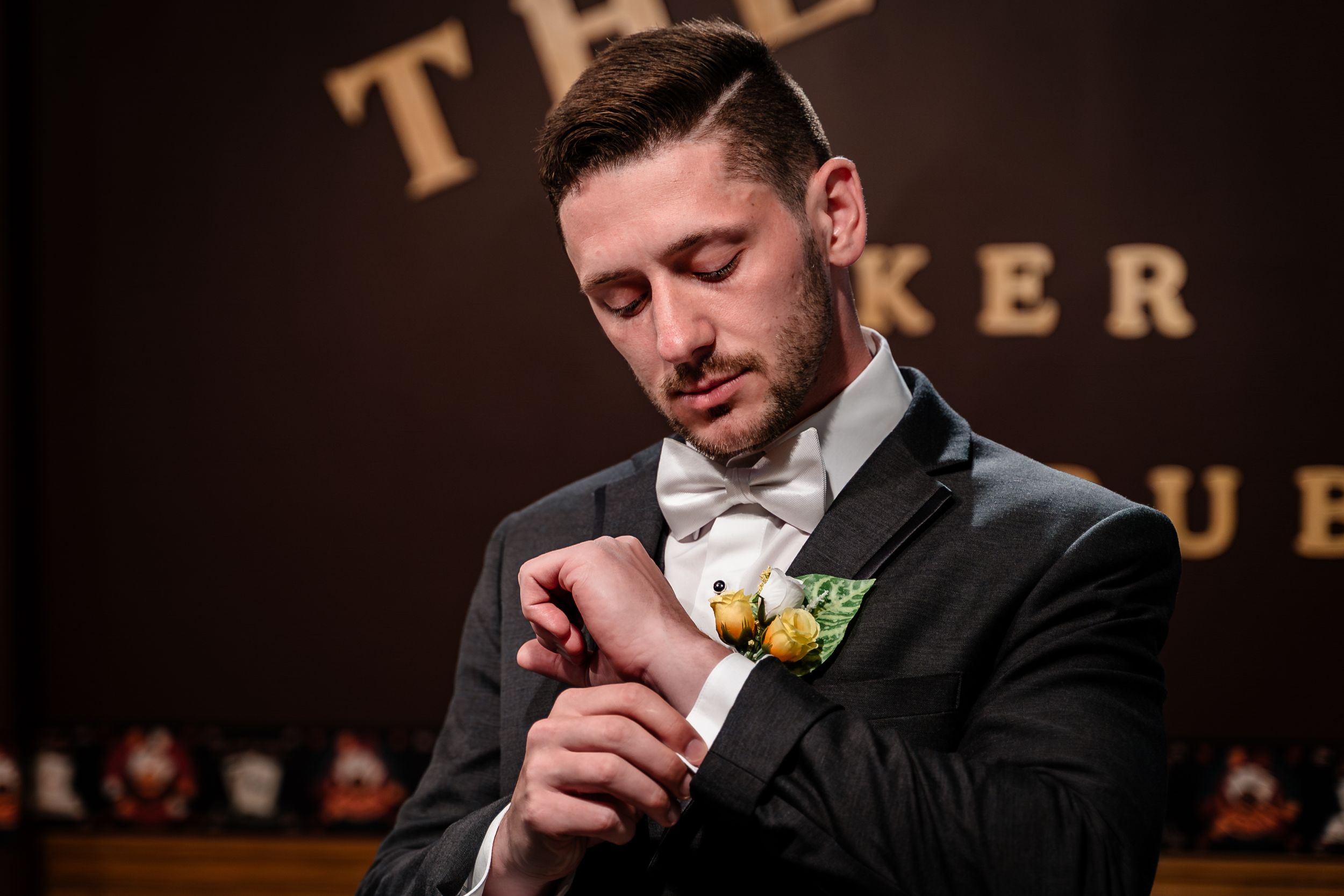 A groom in a black tuxedo and white bowtie checks his watch before the ceremony, with a yellow boutonniere on his lapel.