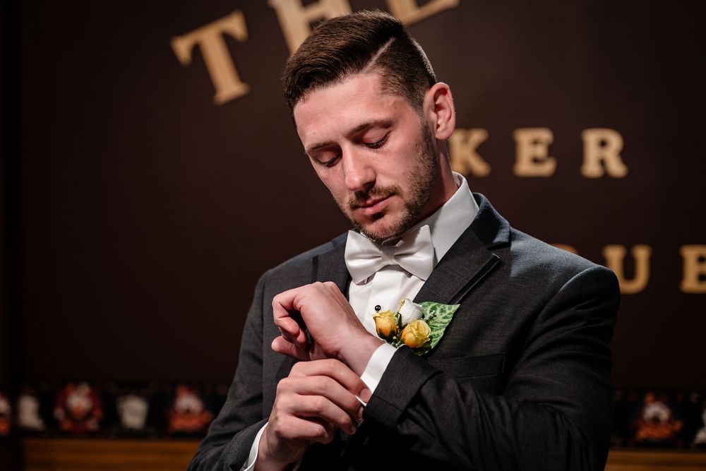 A groom in a black tuxedo and white bowtie checks his watch before the ceremony, with a yellow boutonniere on his lapel.