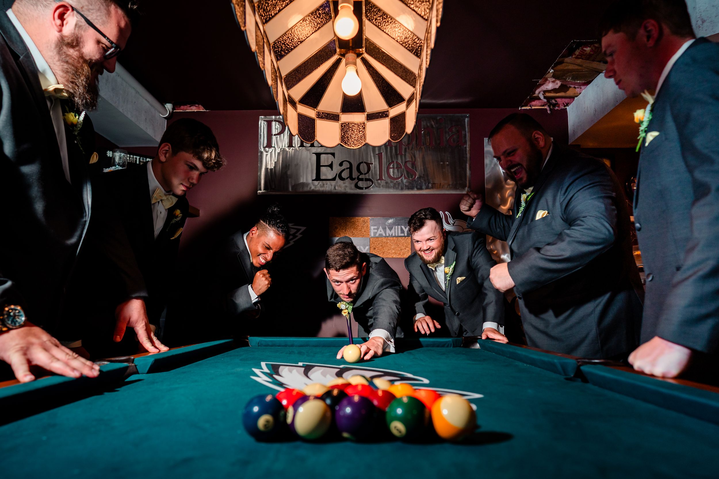 Group of people gathered around a pool table watching billiards game in rustic wooden room with dramatic lighting.