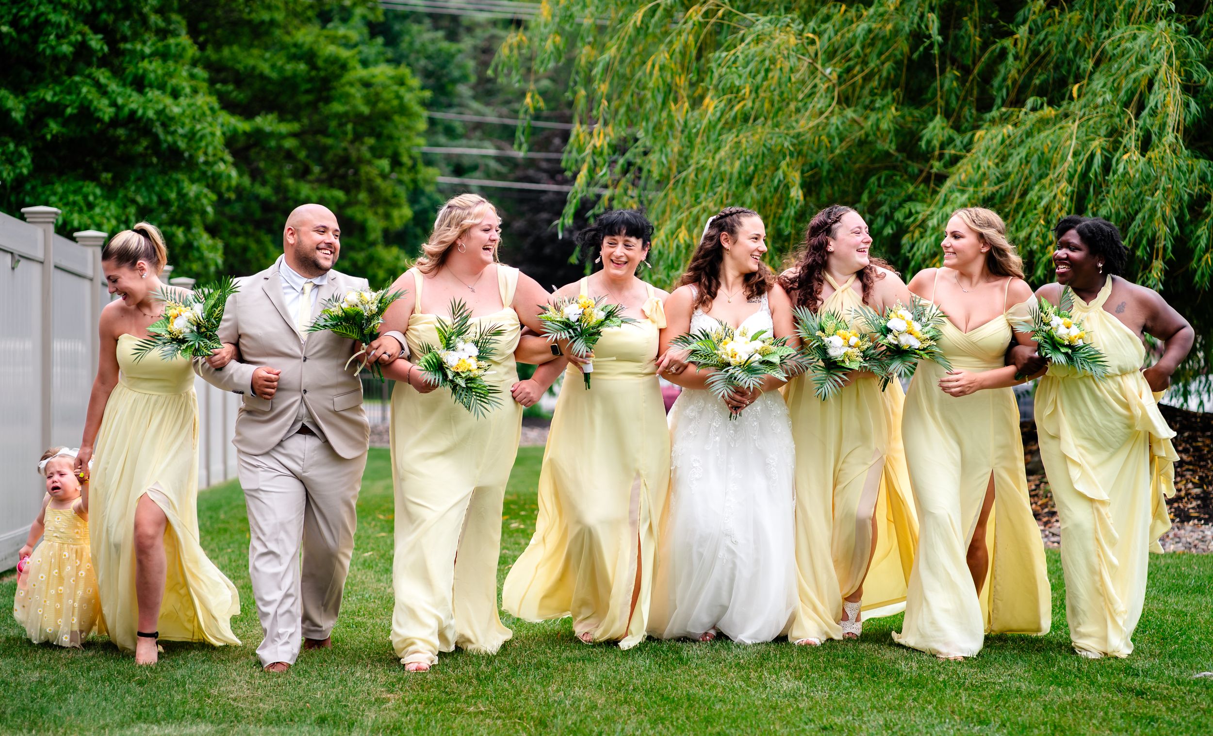 Wedding party wearing yellow and white dresses walk together laughing on green grass during outdoor celebration.