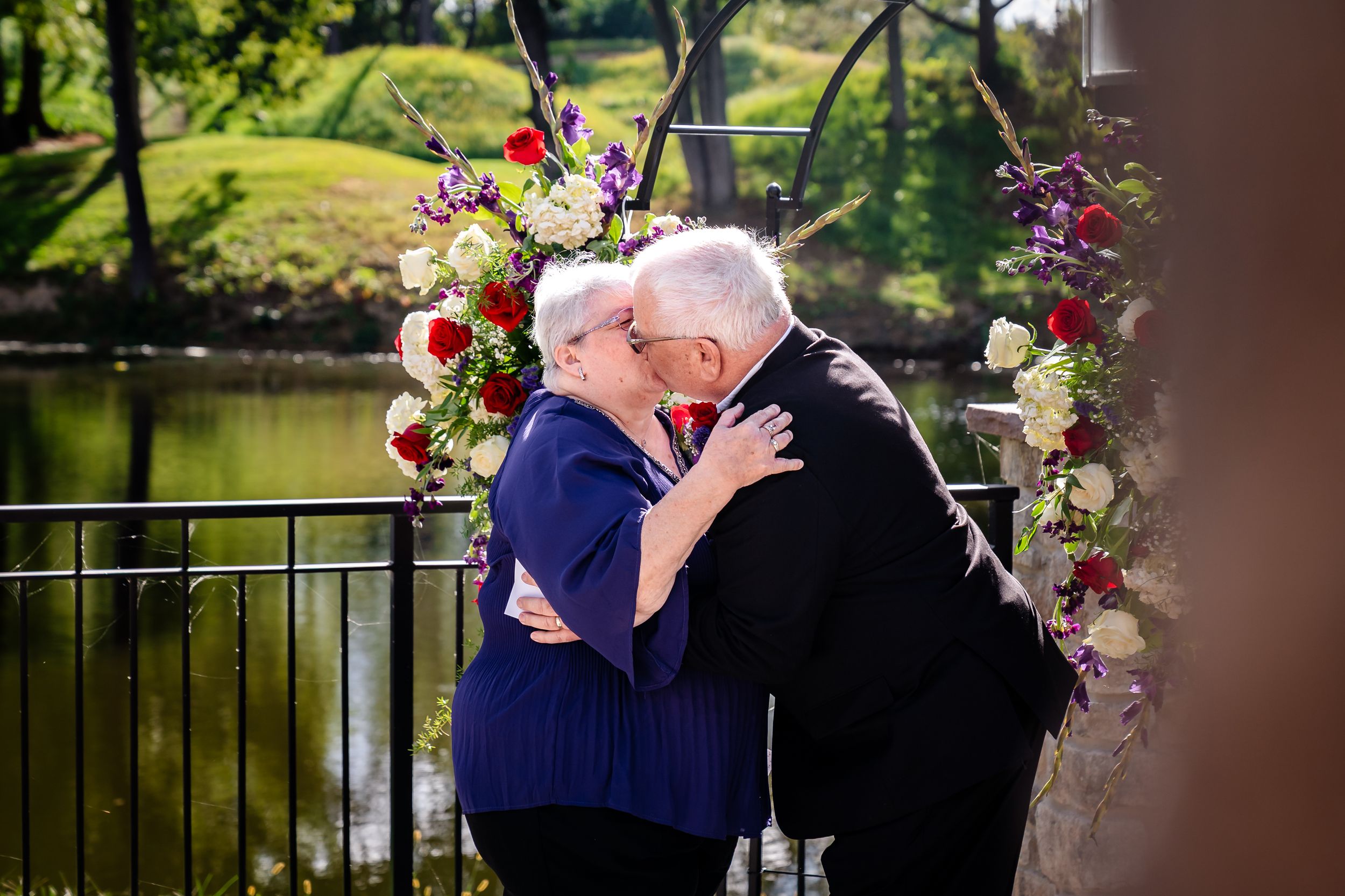 Two people share a tender embrace on a bridge decorated with colorful flowers and floral arrangements near a pond.