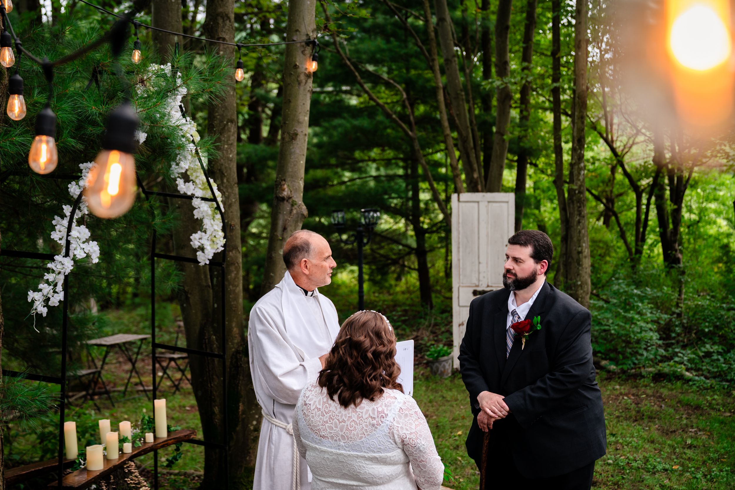 Romantic outdoor wedding ceremony in a wooded setting with glowing string lights and white floral decorations.