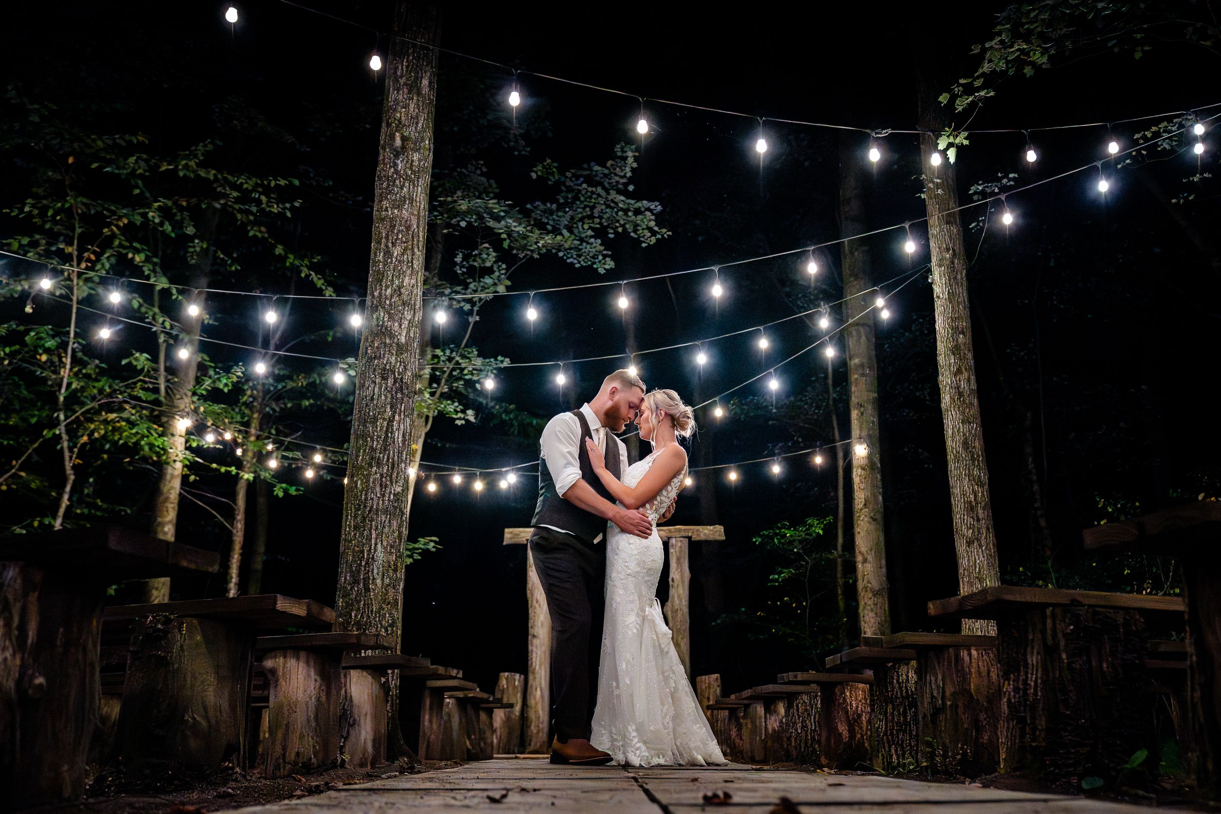 String lights illuminate an outdoor evening wedding reception in a wooded forest setting.
