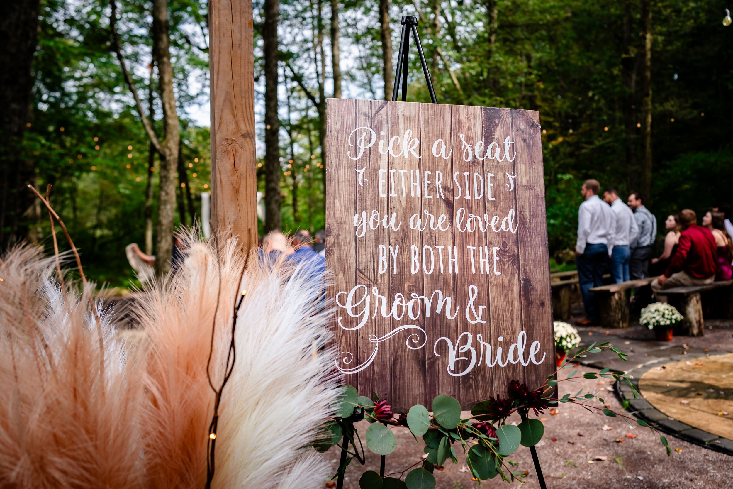 A rustic wooden wedding sign with pampas grass decorations stands in a forest setting during a ceremony.