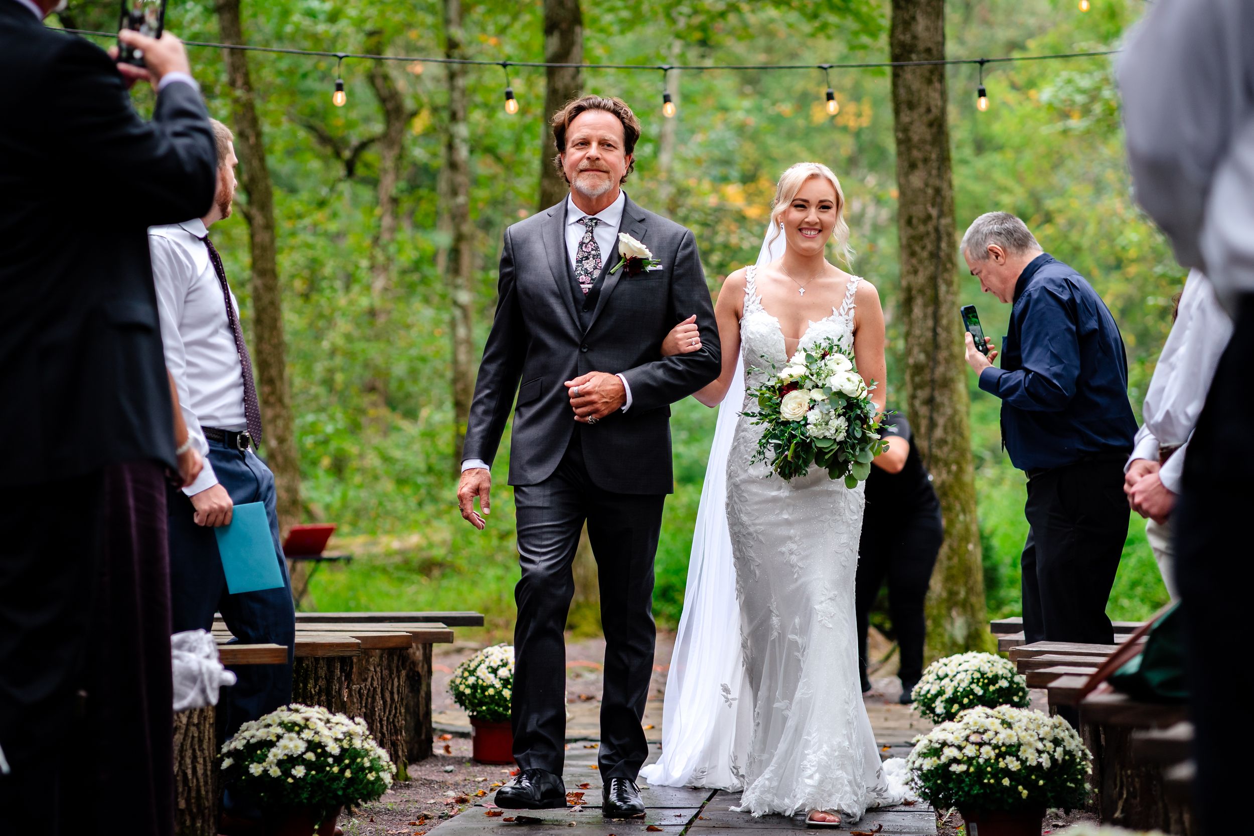 A couple walks down outdoor wooden steps during a forest wedding ceremony surrounded by white floral arrangements.