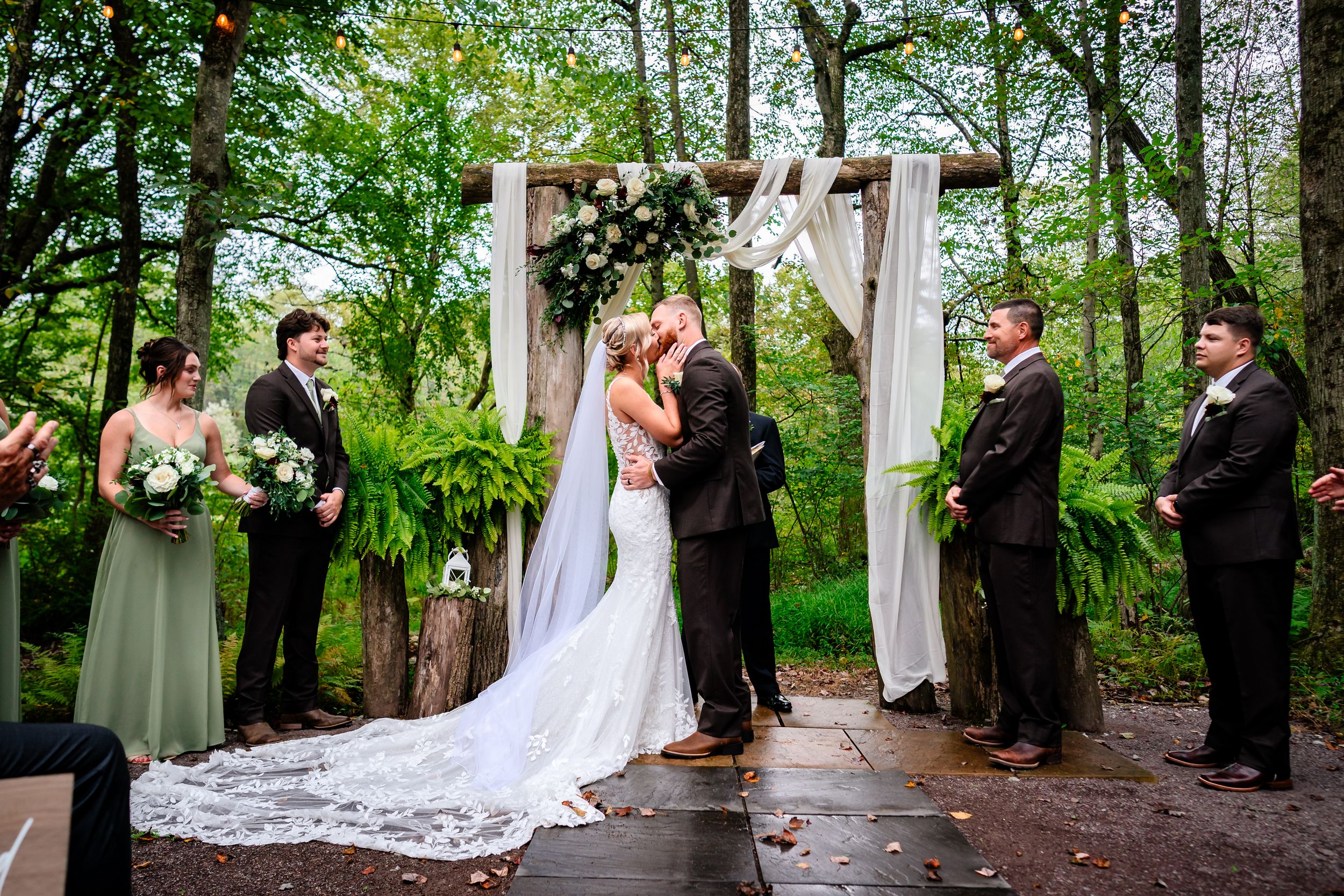 Wedding party stands in a lush green forest setting during an outdoor ceremony.