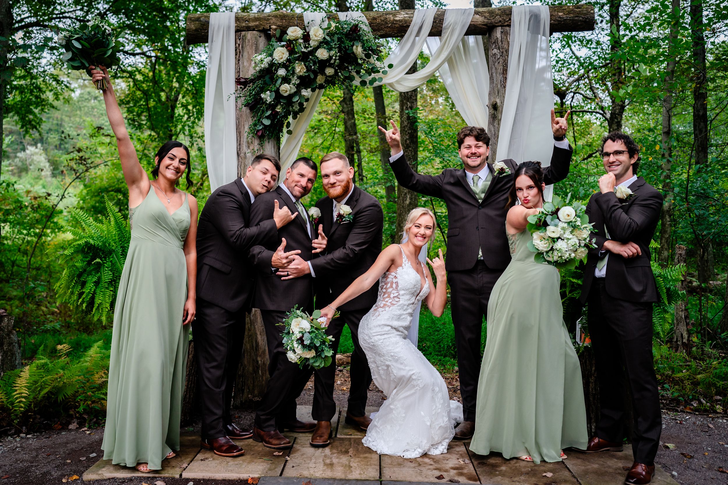 Wedding party poses playfully in a lush garden setting with bridesmaids in sage green dresses.