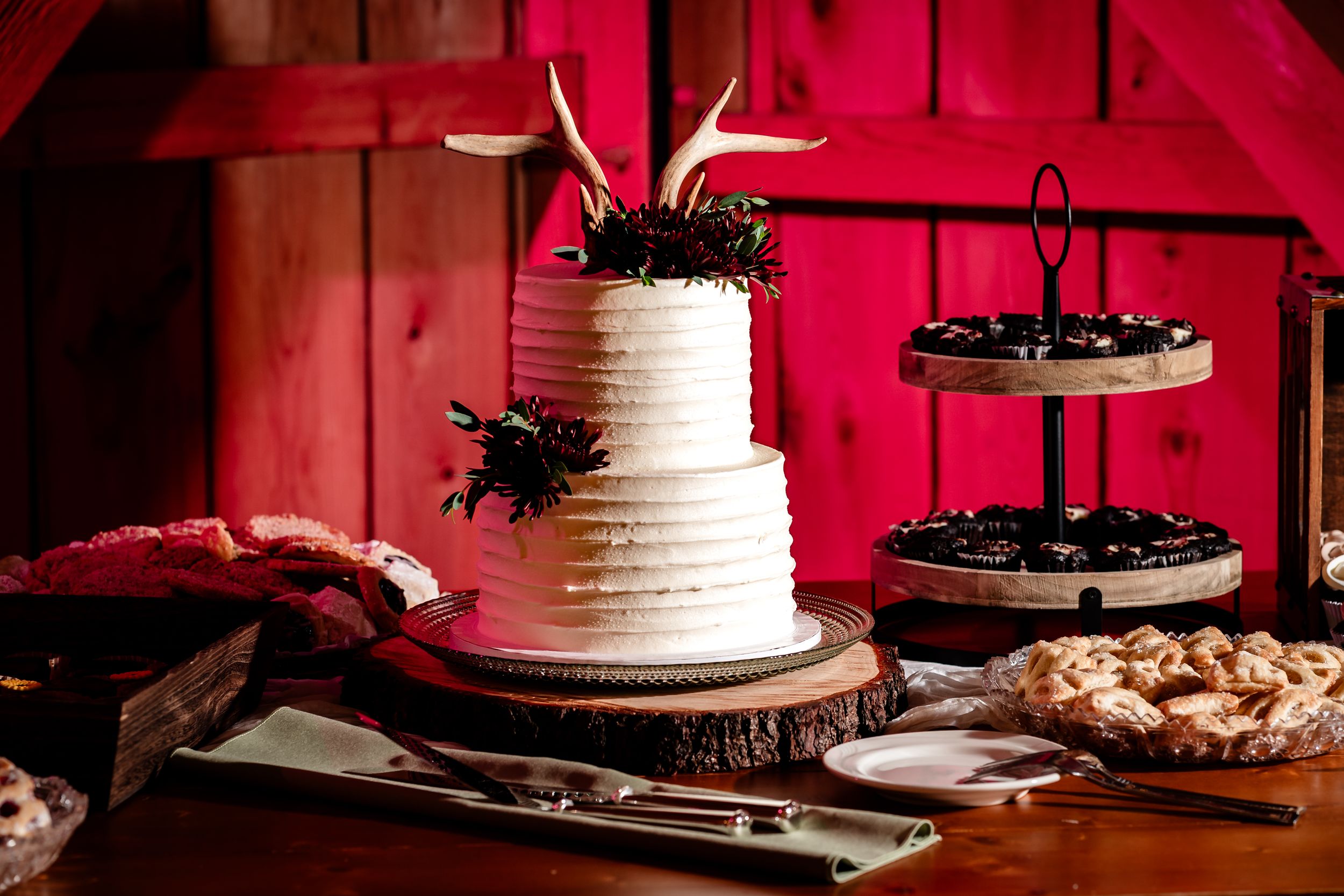 Wedding reception dessert table with white tiered cake and assorted cookies displayed against a dramatic red backdrop.