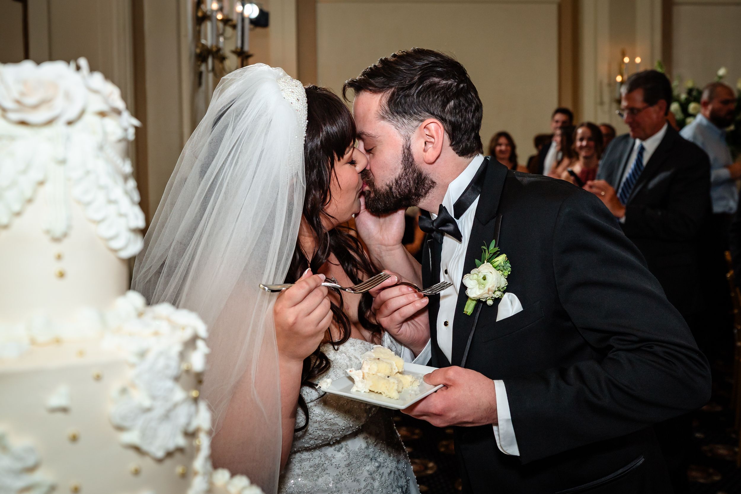 A wedding couple shares a romantic kiss during their reception next to an elegant white wedding cake.