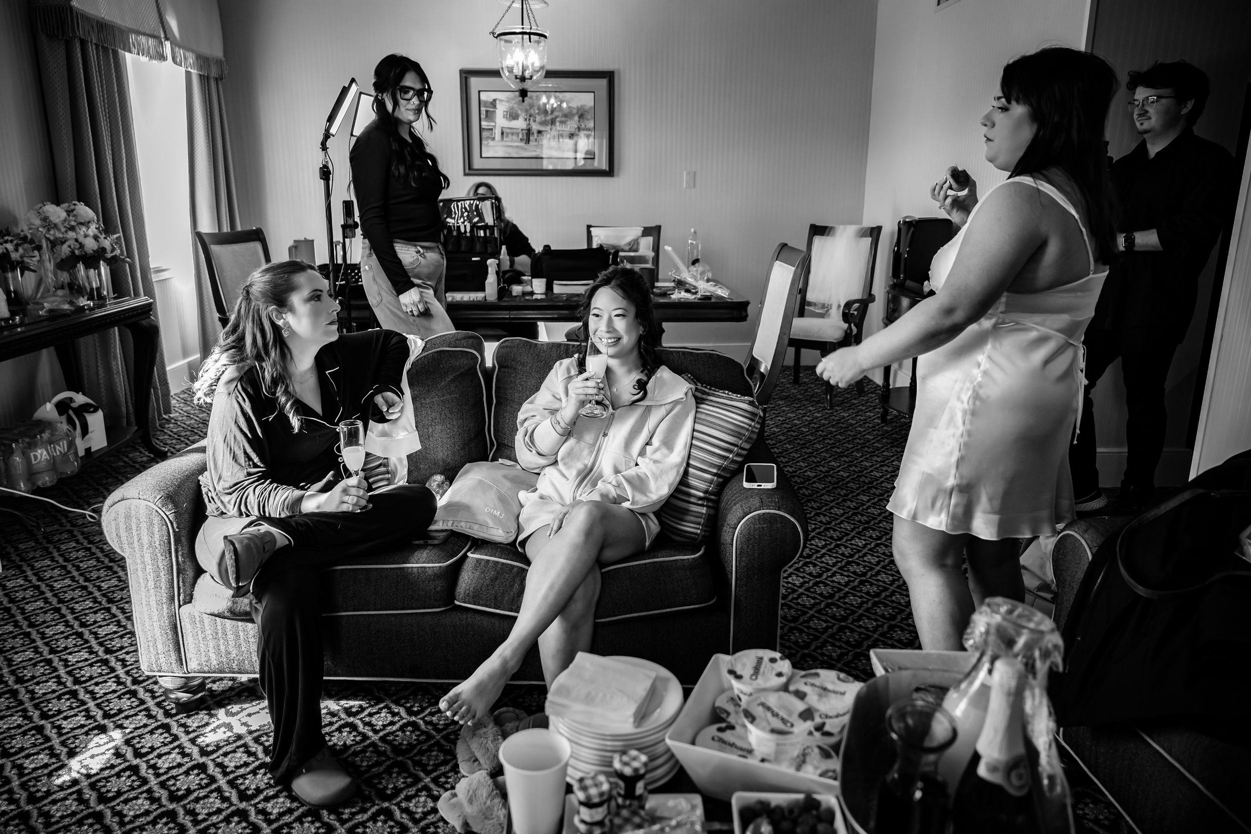 A candid black and white photograph showing people relaxing in a living room during wedding preparations.