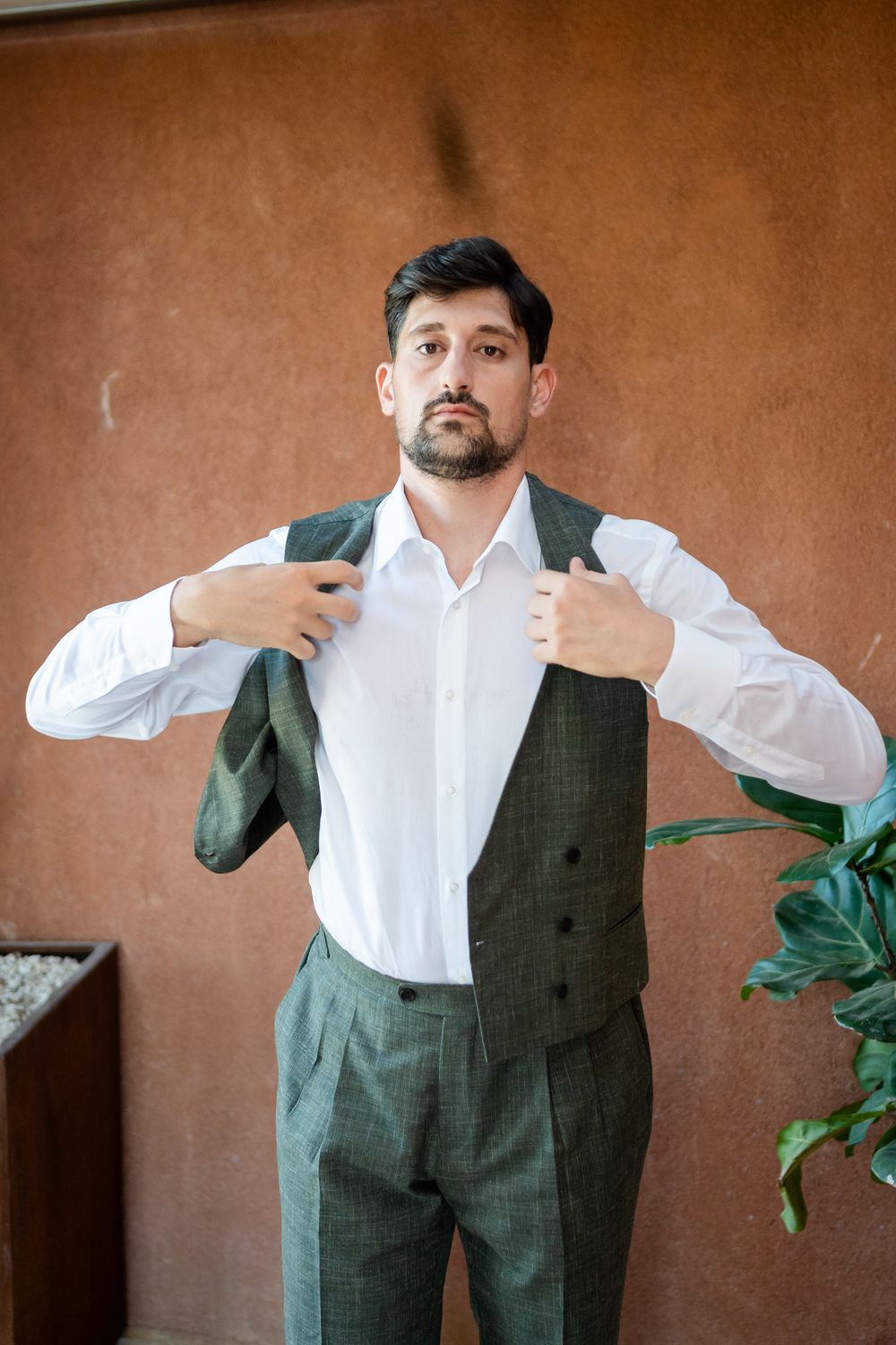 A person wearing a formal olive green suit vest and white dress shirt stands against a terracotta colored wall.