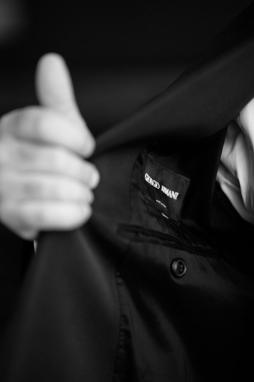 A black and white close-up photo showing a thumbs up gesture and a dark jacket with visible clothing tag.
