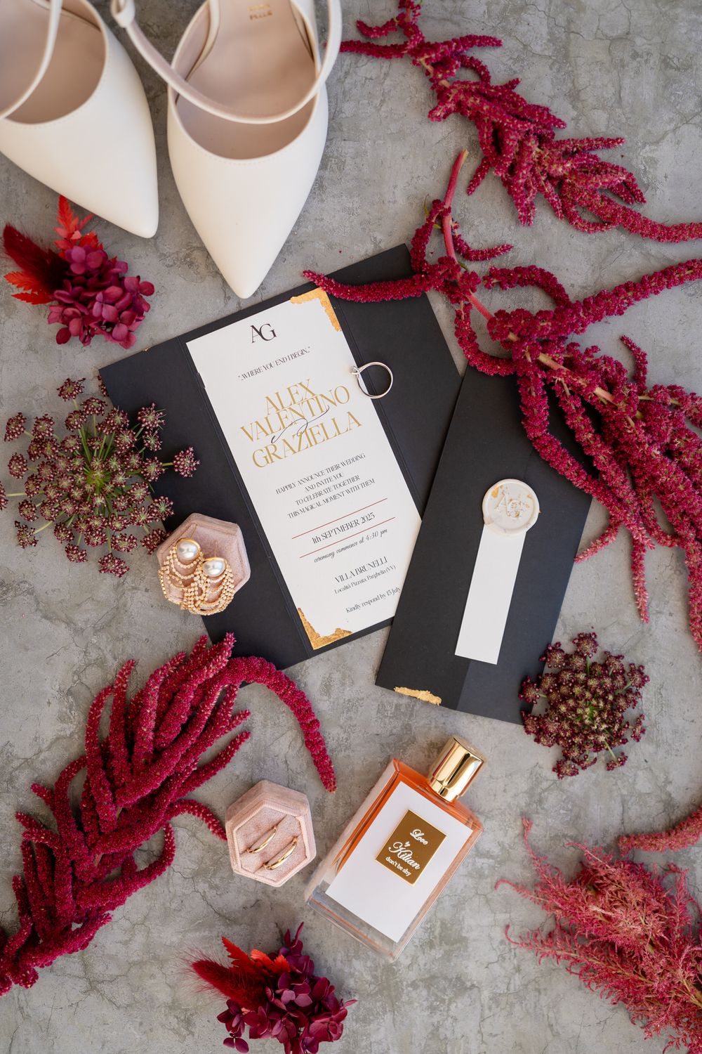 Wedding details flatlay with white shoes, red amaranth flowers, perfume bottles and invitation cards on grey surface.