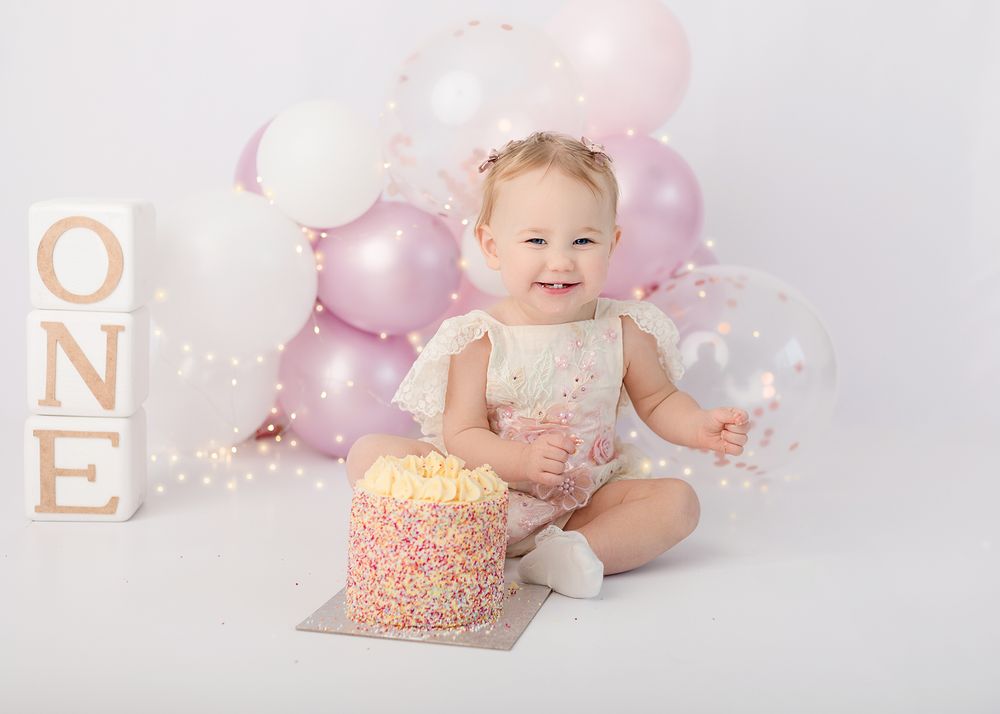 Baby in white outfit sits with pink and white balloons and smash cake during first birthday photo session.