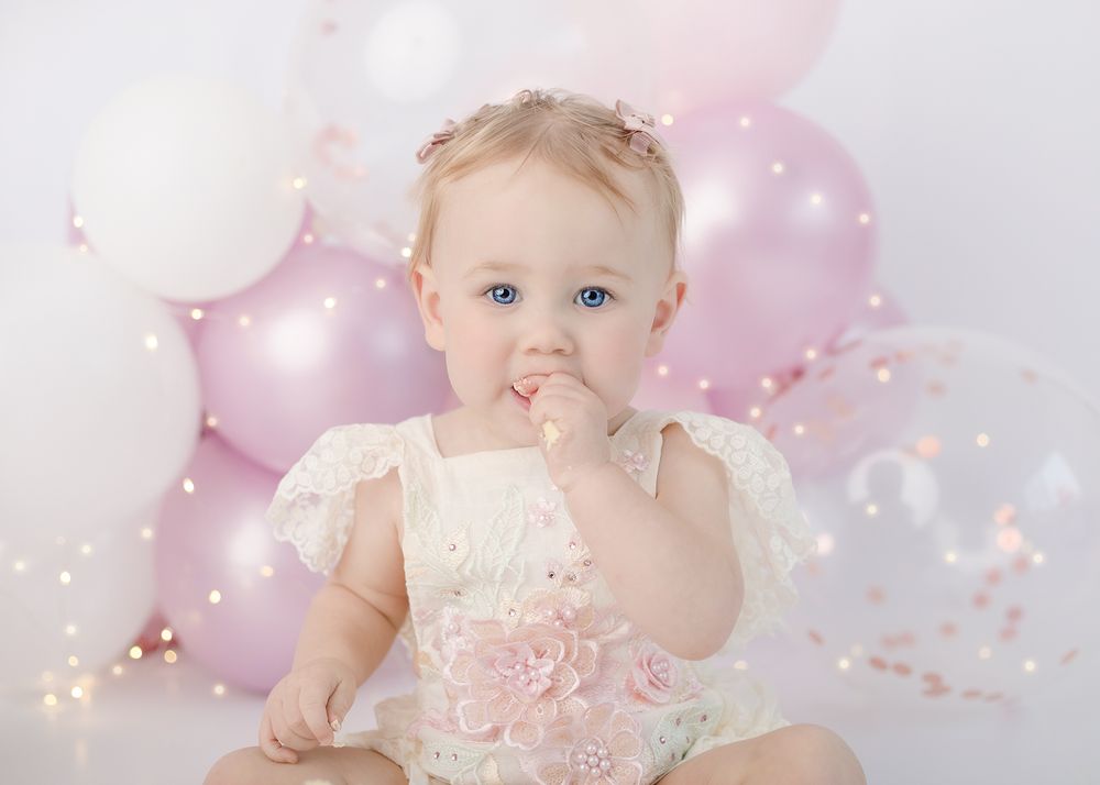Baby in pink dress sits among pink and white balloons with confetti during first birthday cake smash photo session.
