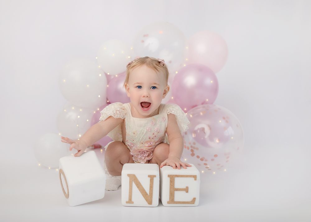 Baby crawling on white blanket with pink balloons and gold ONE letters for first birthday photoshoot.