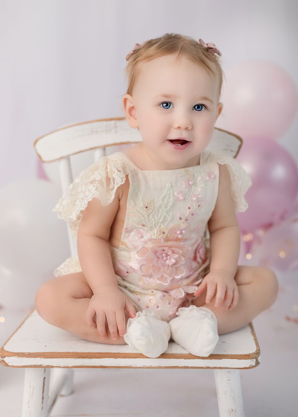 Baby in delicate cream lace dress sits on white wooden chair with pink balloons and soft lighting in background.